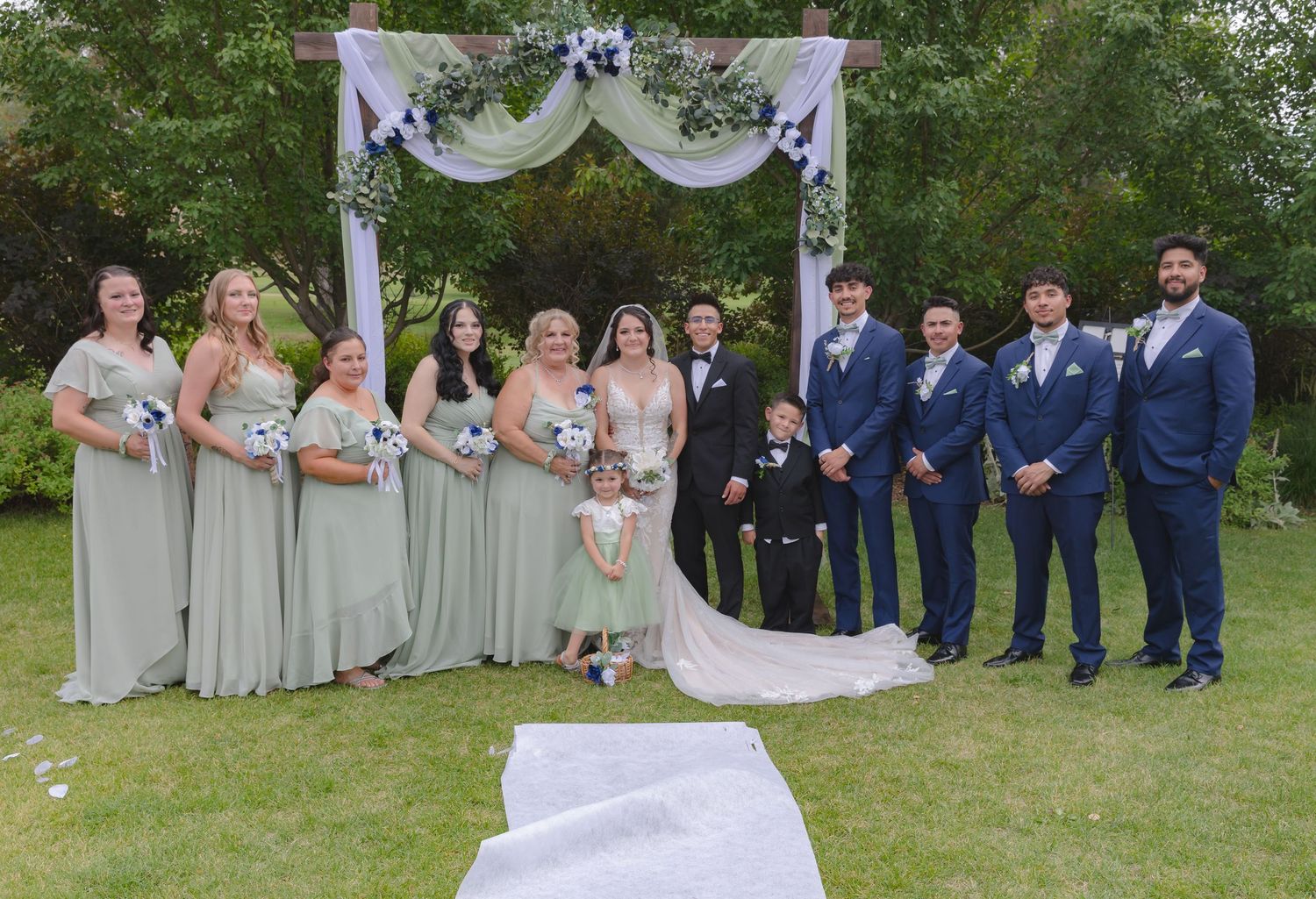 Outdoor wedding ceremony with wedding party in gray and navy blue attire posed under decorated archway on grass.