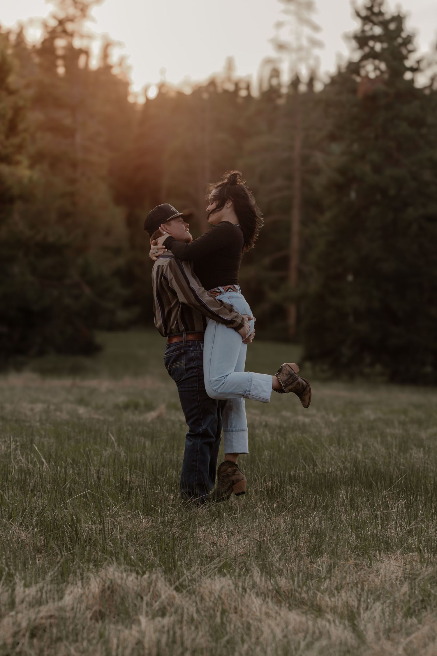 Romantic outdoor portrait of couple hugging in field during sunset with forest backdrop.