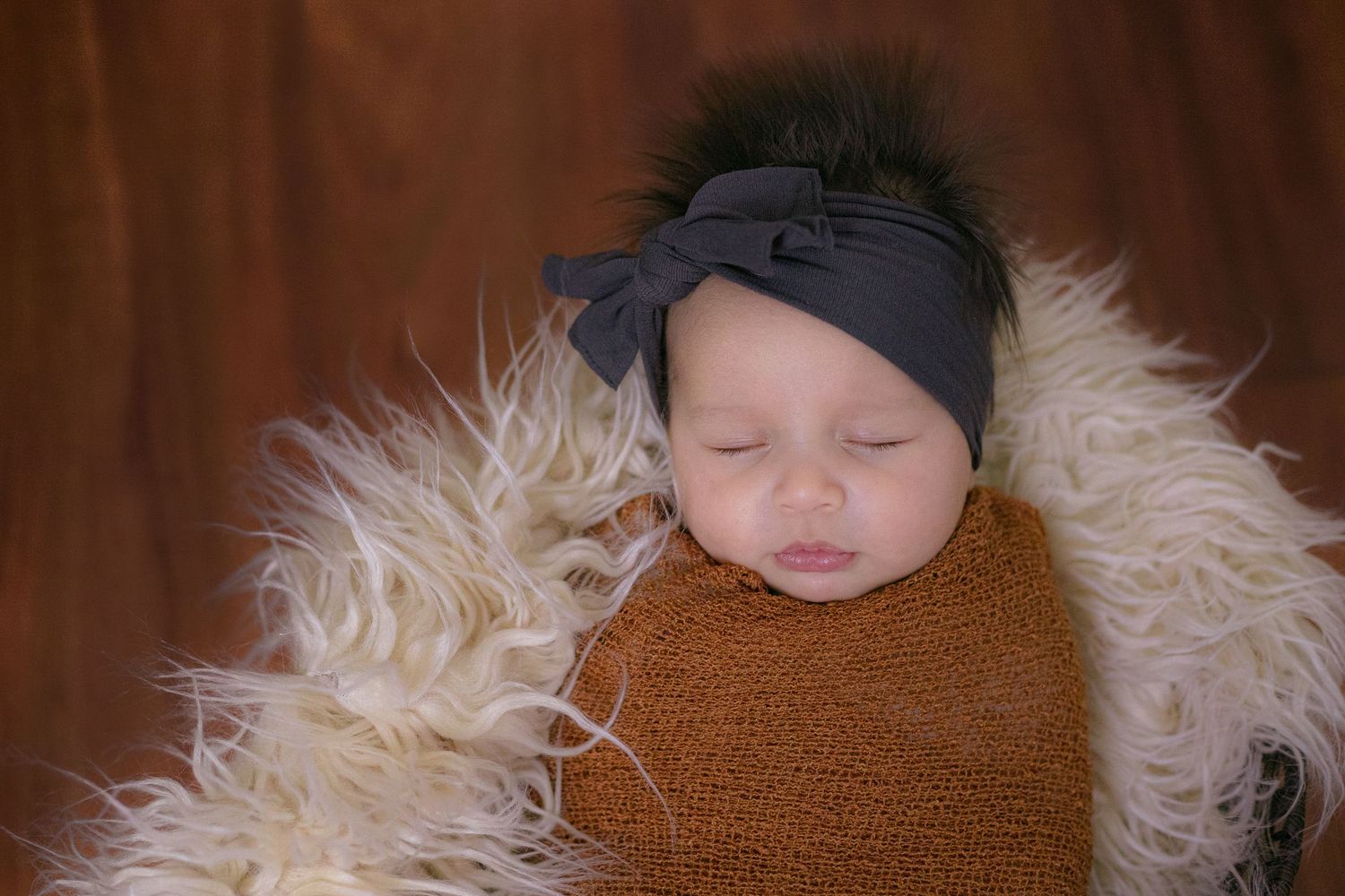 Sleeping baby wrapped in brown swaddle blanket rests on fluffy white fur with dark grey headband.
