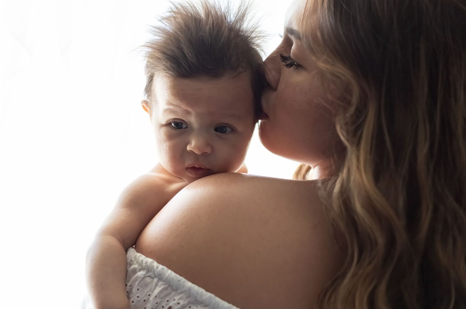 Mother tenderly kisses and holds baby in warm sunlit room creating an intimate bonding moment.