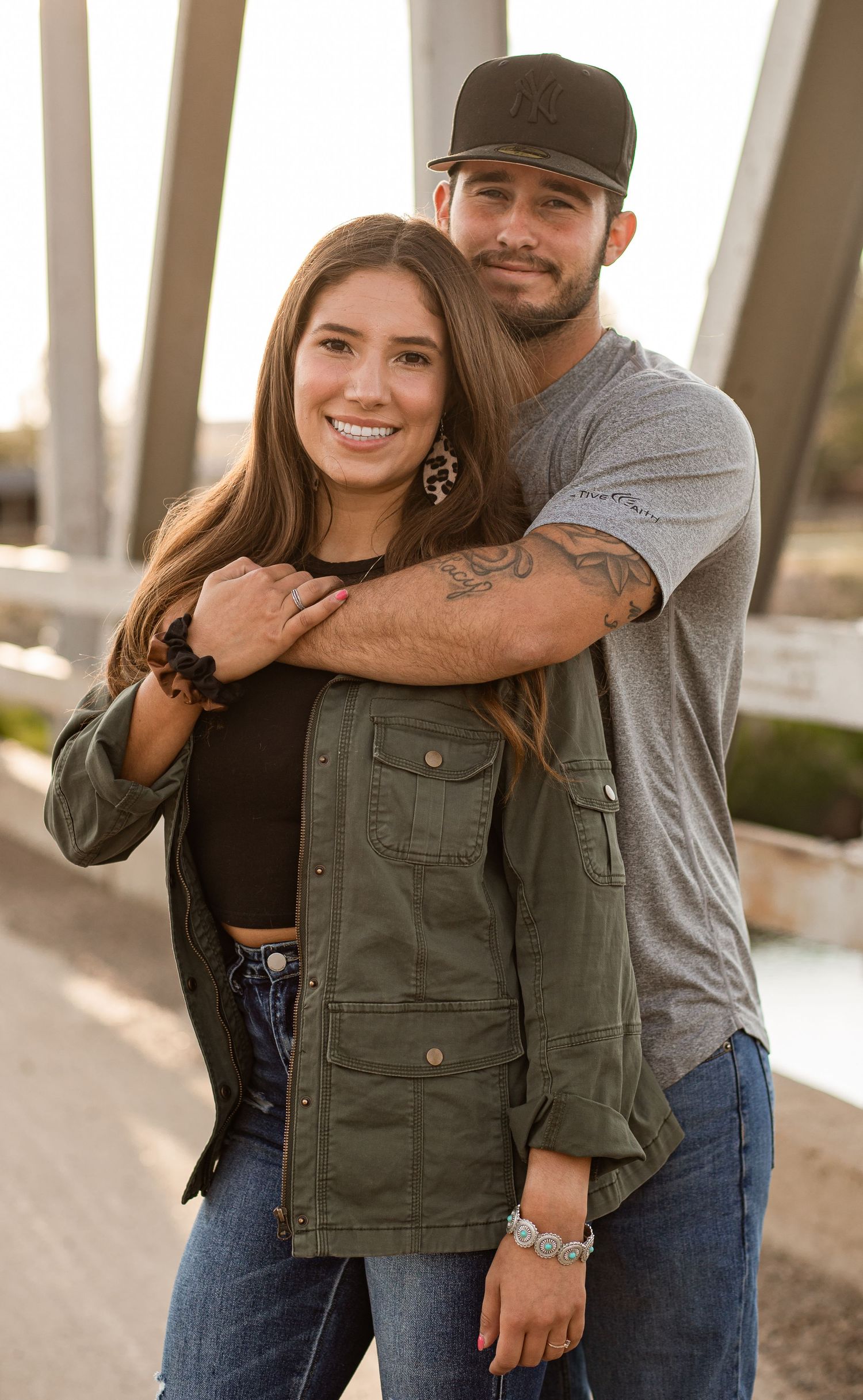 A couple in casual attire embracing and smiling outdoors, wearing jeans and jackets against a soft-focus background.