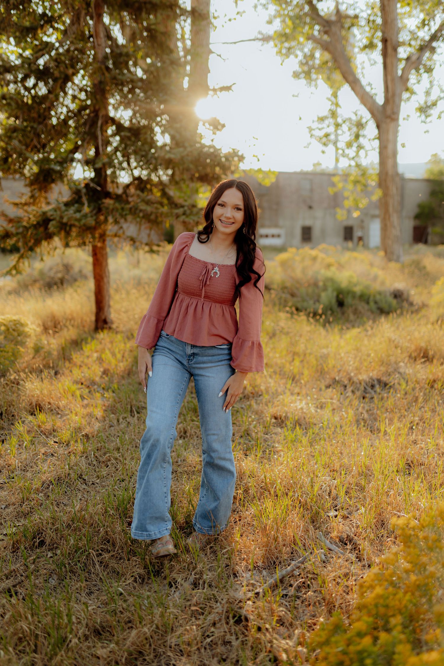 Young woman in pink top and flared jeans poses outdoors in golden sunlit field with trees.