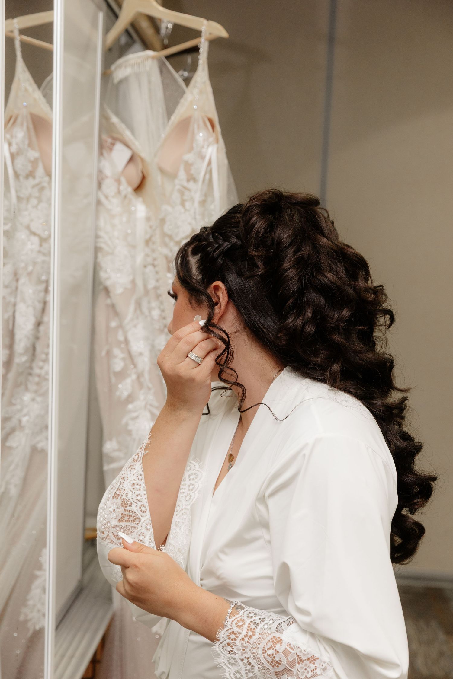 Bride in white robe looks at hanging wedding dress while getting ready with elegant curled hairstyle.
