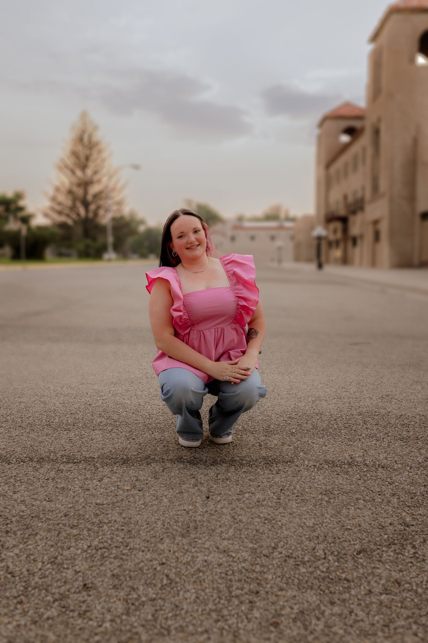 Woman in pink ruffled top and jeans crouching on pavement, smiling outdoors near buildings at dusk.