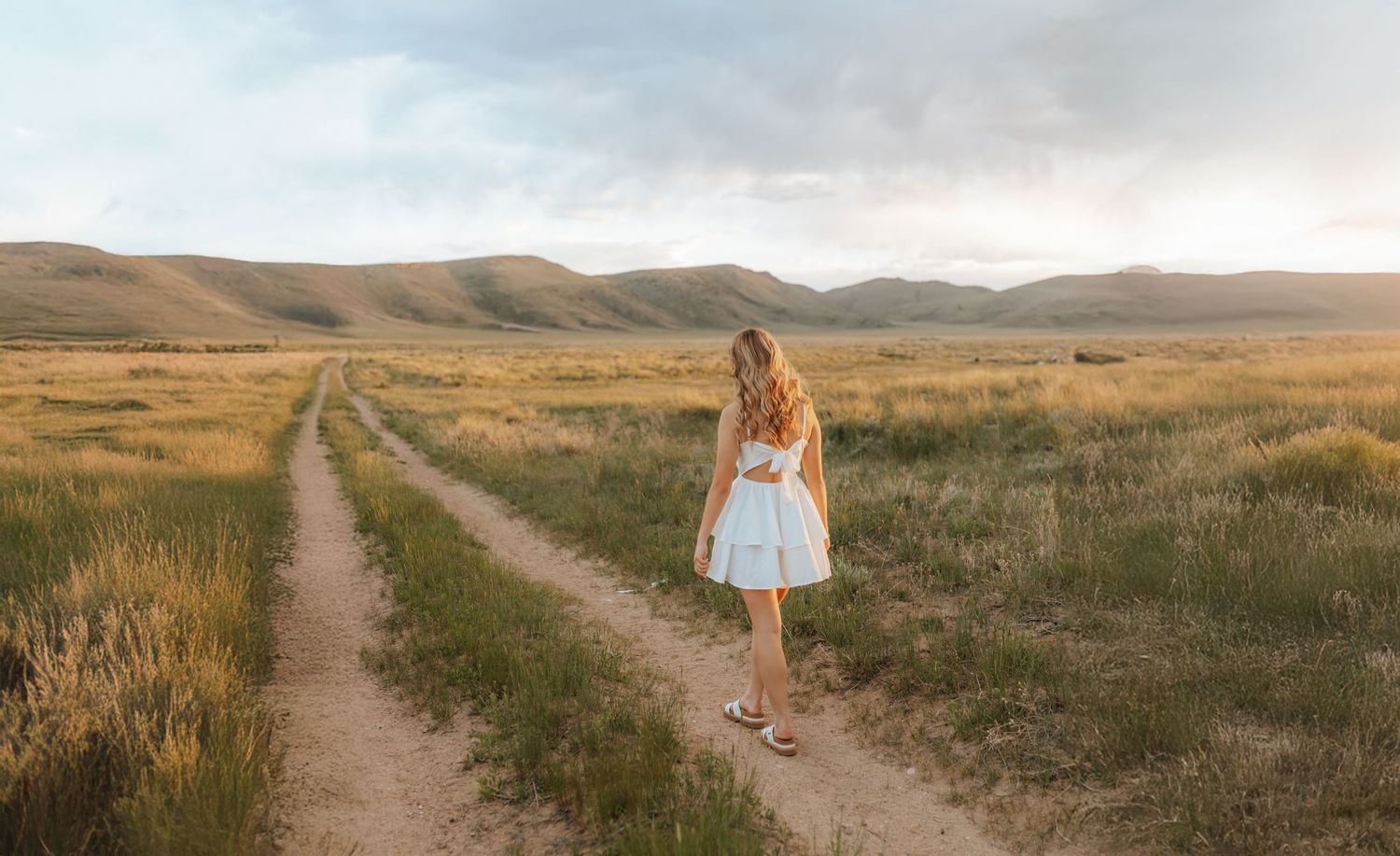 A person in a white dress walking down a dirt road through rolling hills.