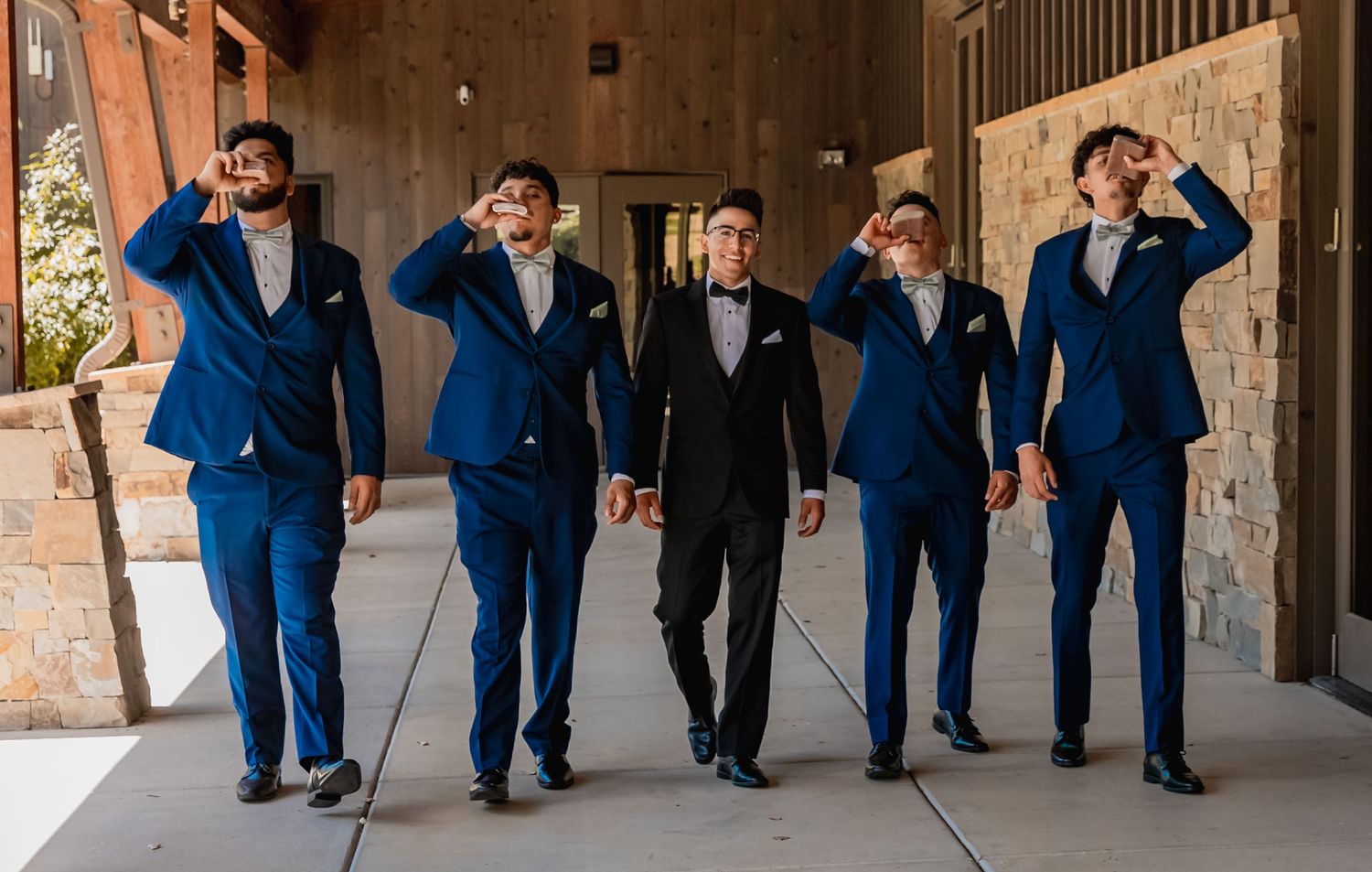 Groomsmen in matching navy blue suits walk and pose playfully in a rustic covered walkway setting.