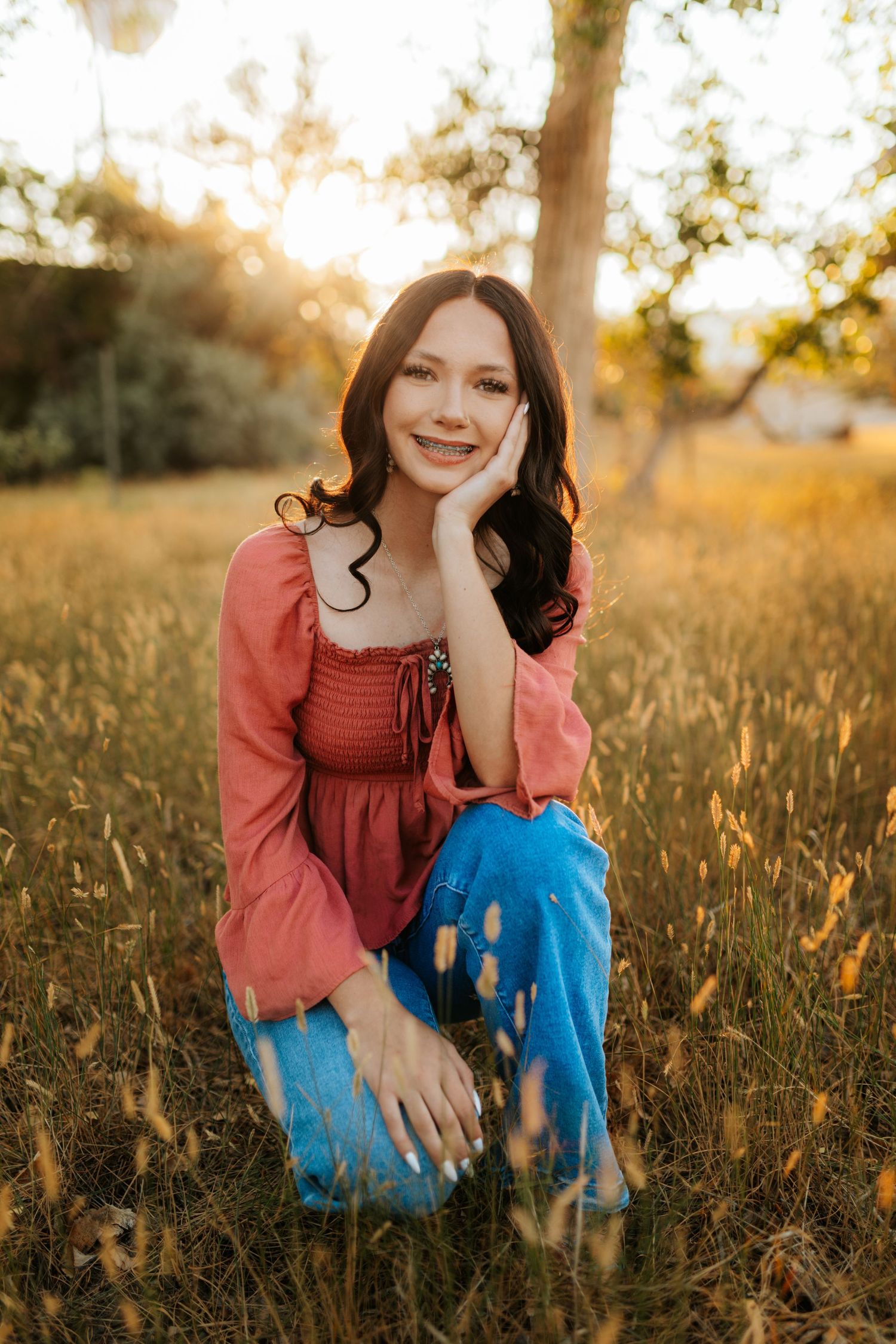 Young woman in coral top and blue jeans sitting in golden field at sunset, smiling with hand on cheek.