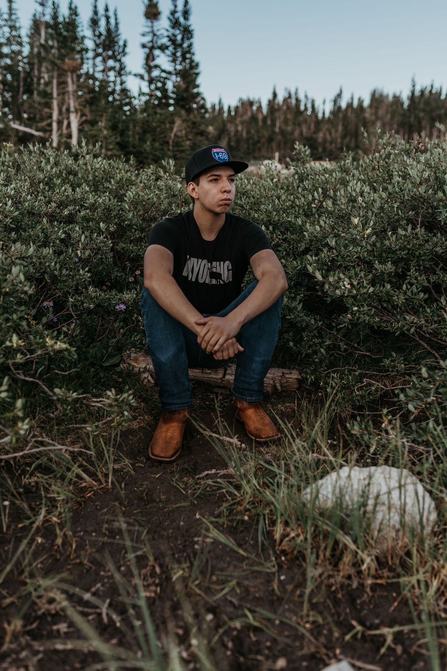 Young man in Wyoming shirt and cap sitting on log surrounded by wild shrubs and pine trees outdoors.