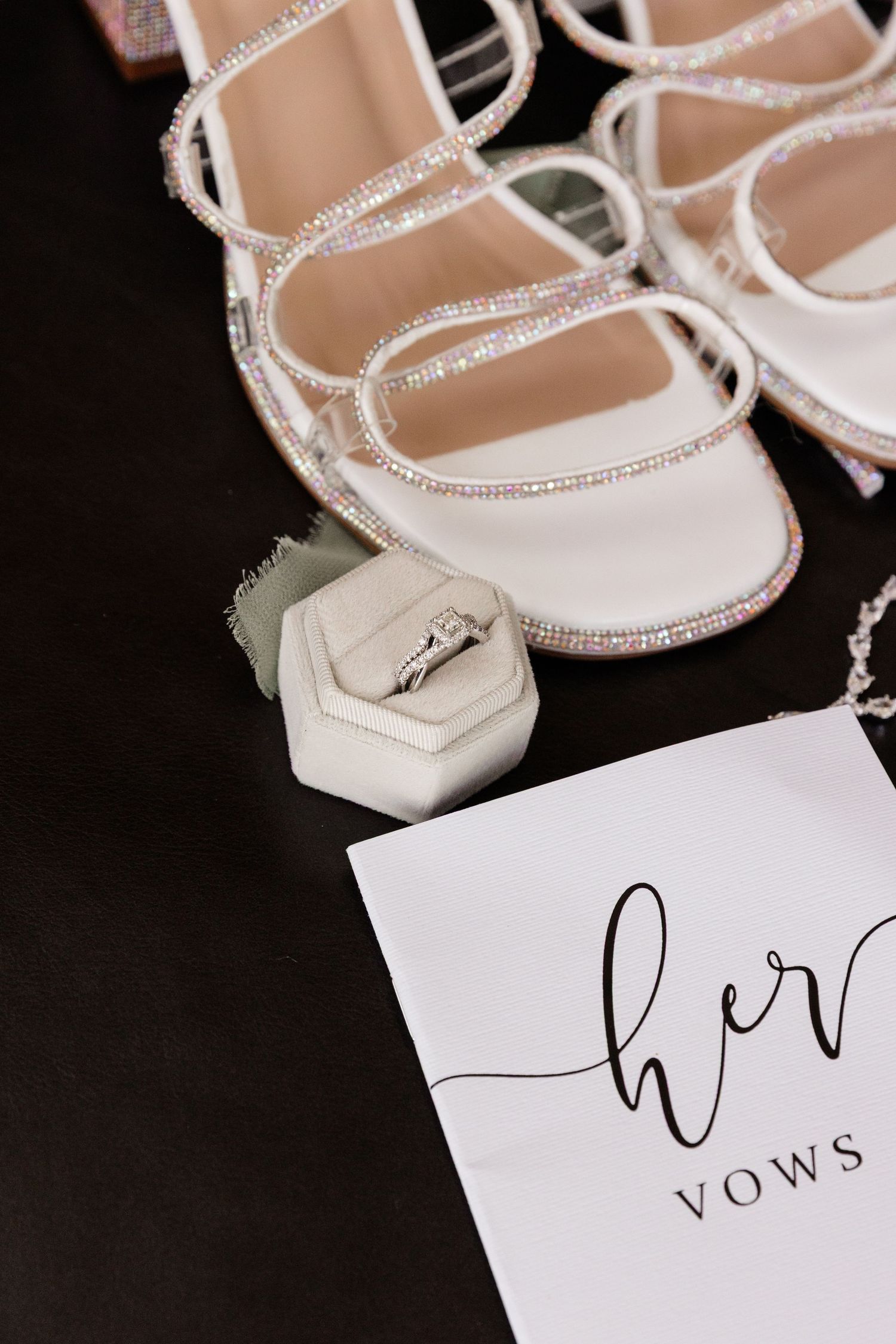 White crystal-embellished bridal sandals next to a hexagonal ring box and vows card on a black surface.