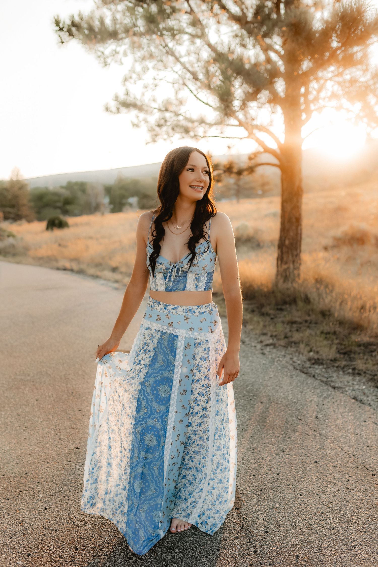 Young woman in blue floral two-piece outfit standing on a sunlit road at golden hour.
