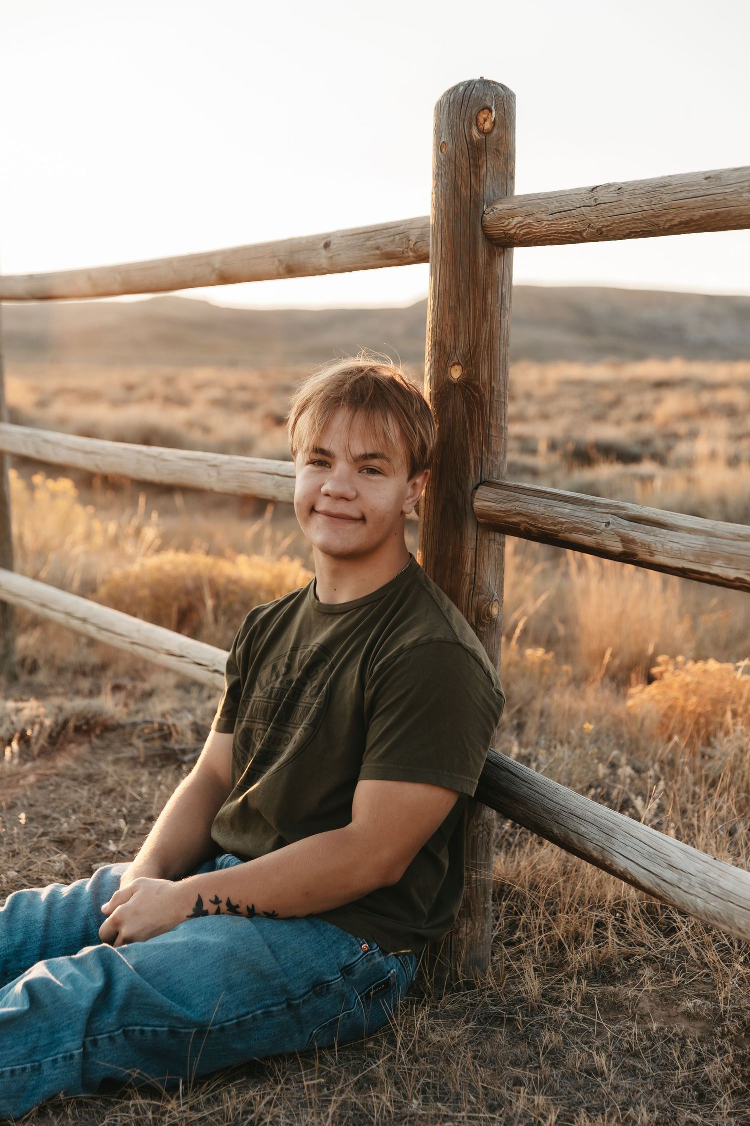 A smiling teenage boy in a green t-shirt and jeans sits against a rustic wooden fence in a golden field at sunset.
