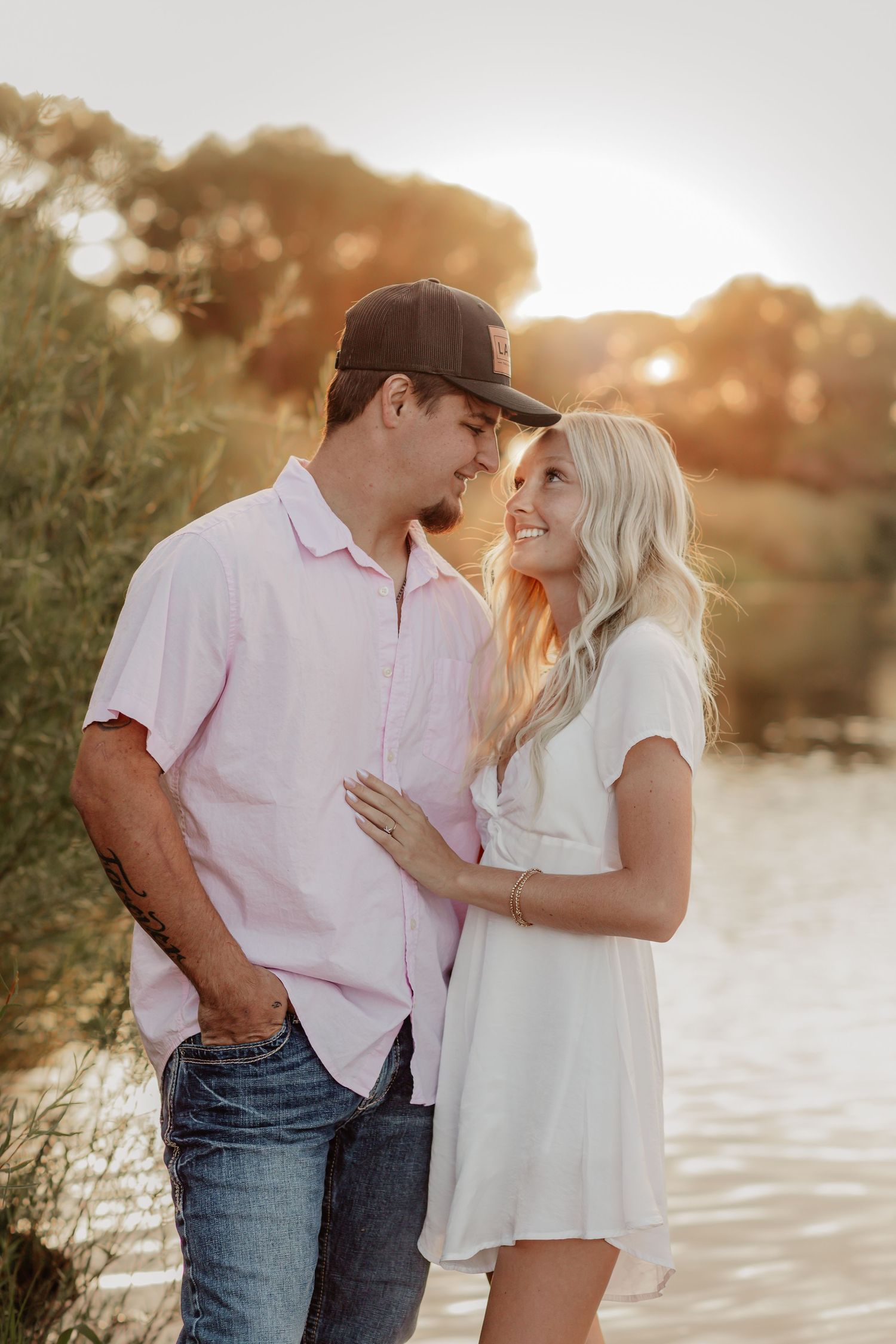 Romantic couple embraces at sunset by the water's edge with warm golden light glowing through trees.
