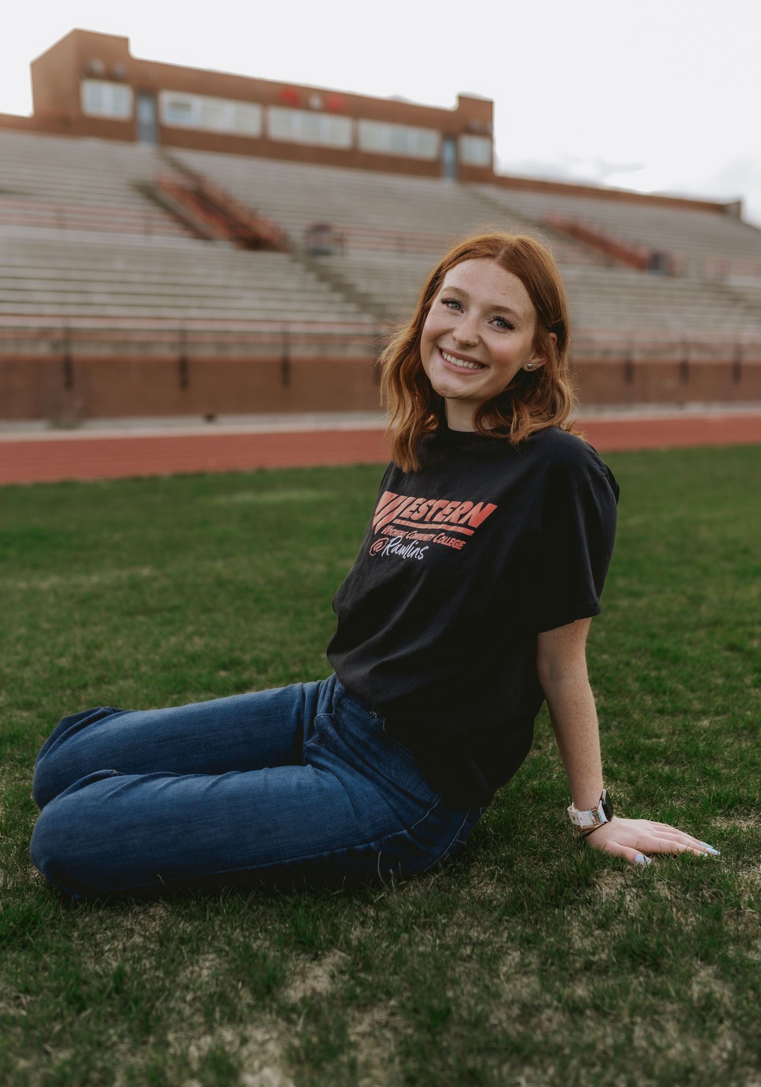 Smiling redhead woman sitting on grass in a black university t-shirt and jeans near a stadium track.
