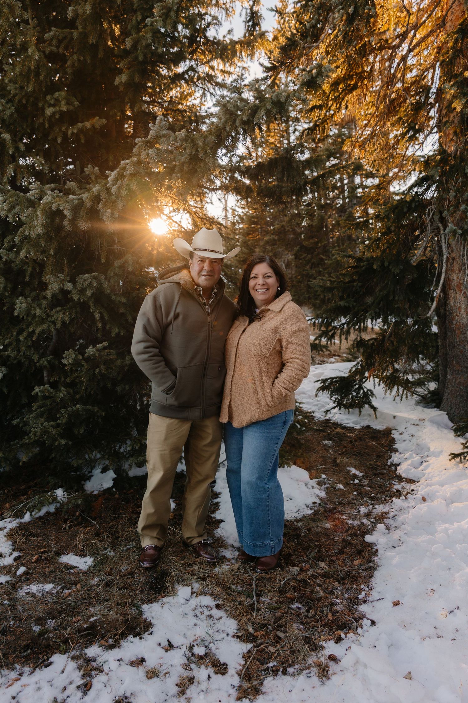 Couple smiling together among snow-dusted evergreen trees at golden sunset in a winter forest setting.