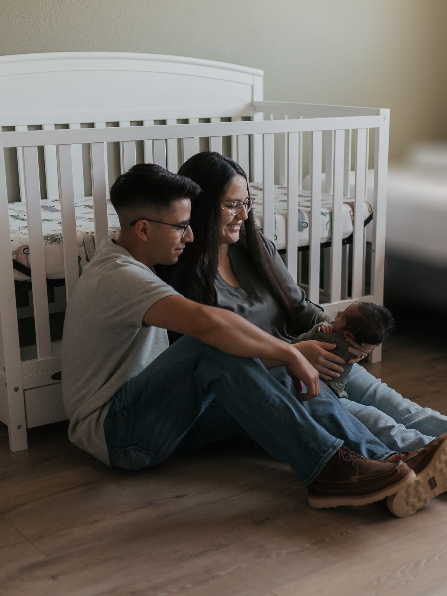 A couple sits together on the floor against a white crib, sharing tender moments and looking at their phones together.