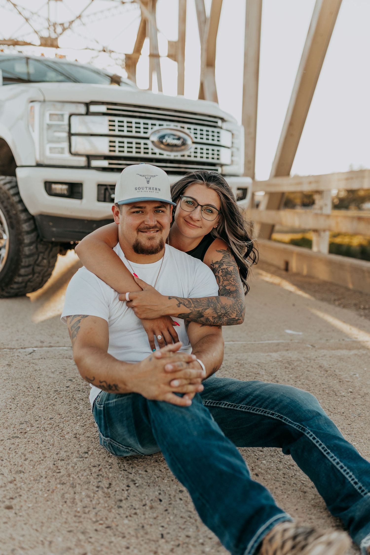 Couple posing on a bridge in front of a white Ford truck, woman hugging man from behind, both wearing jeans.
