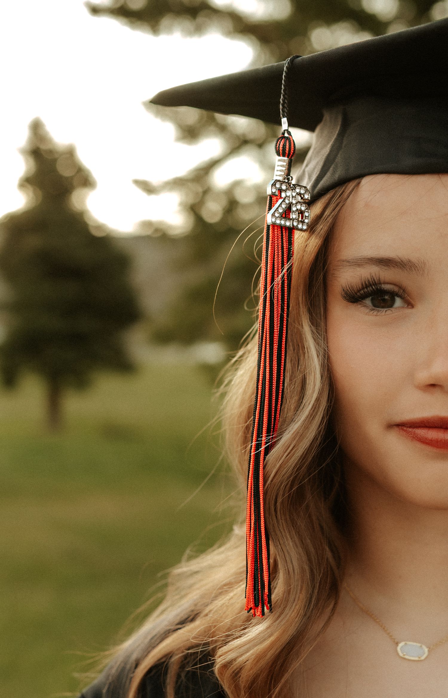 Young woman in black graduation cap with red tassel smiles outdoors, celebrating her academic achievement.