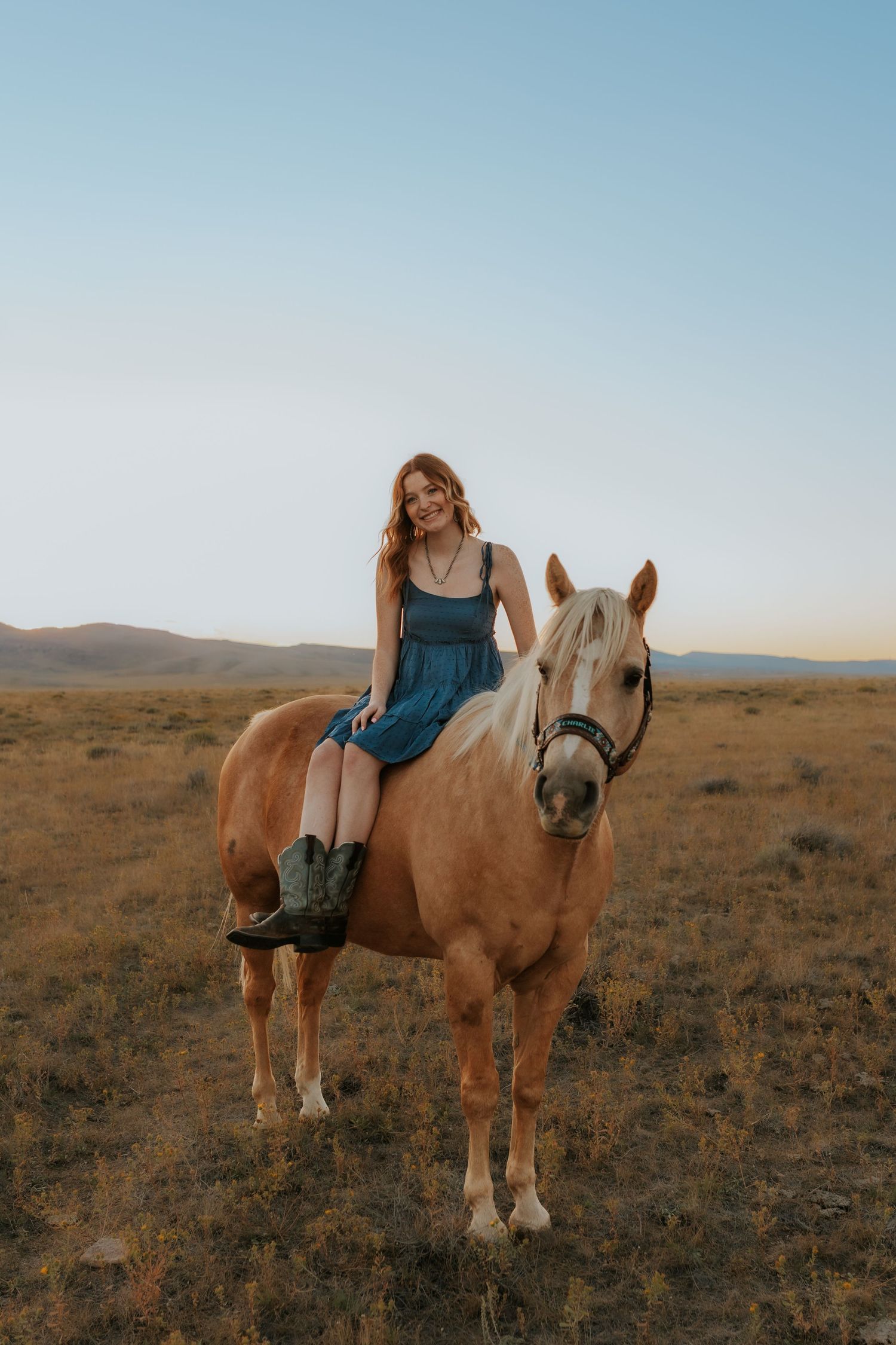 A young woman in a blue dress and cowboy boots rides a palomino horse in an open field at dusk.