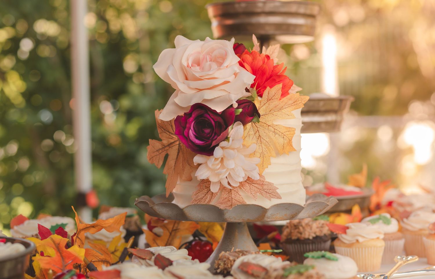 A delicate wedding cake adorned with pink and red roses sits among autumn leaves during golden hour.