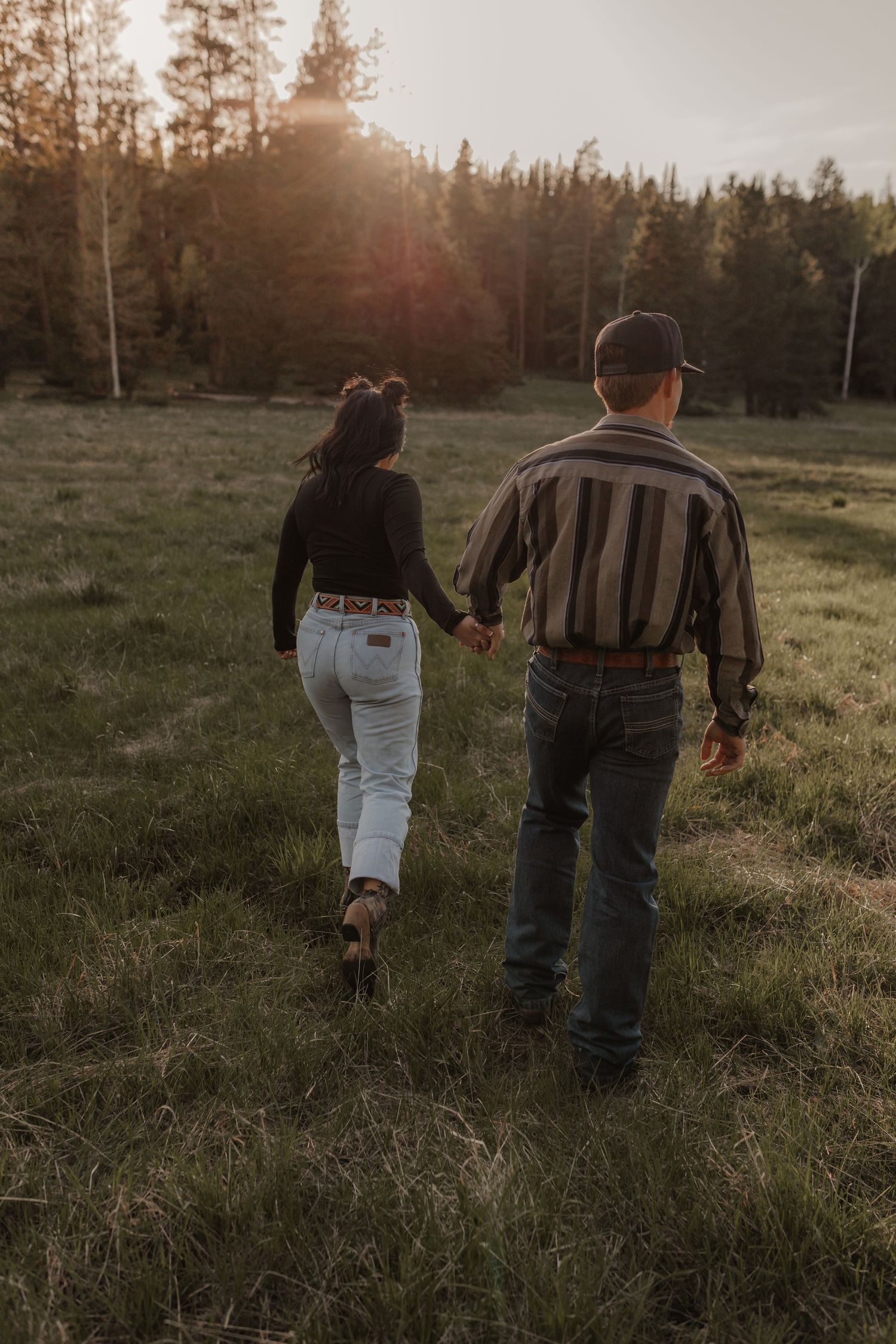 Two people walking hand in hand through a grassy field at sunset with pine trees in background.