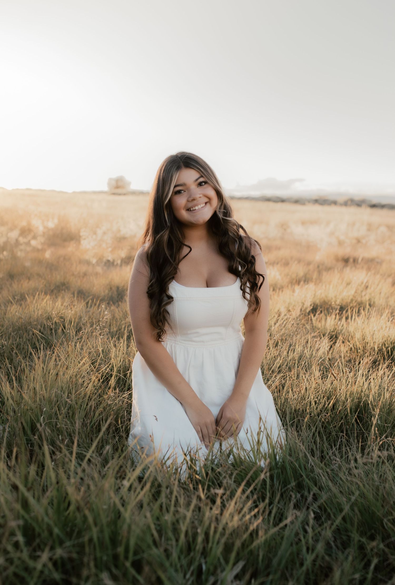 Young woman in white dress sitting in golden field at sunset, smiling with long dark wavy hair.