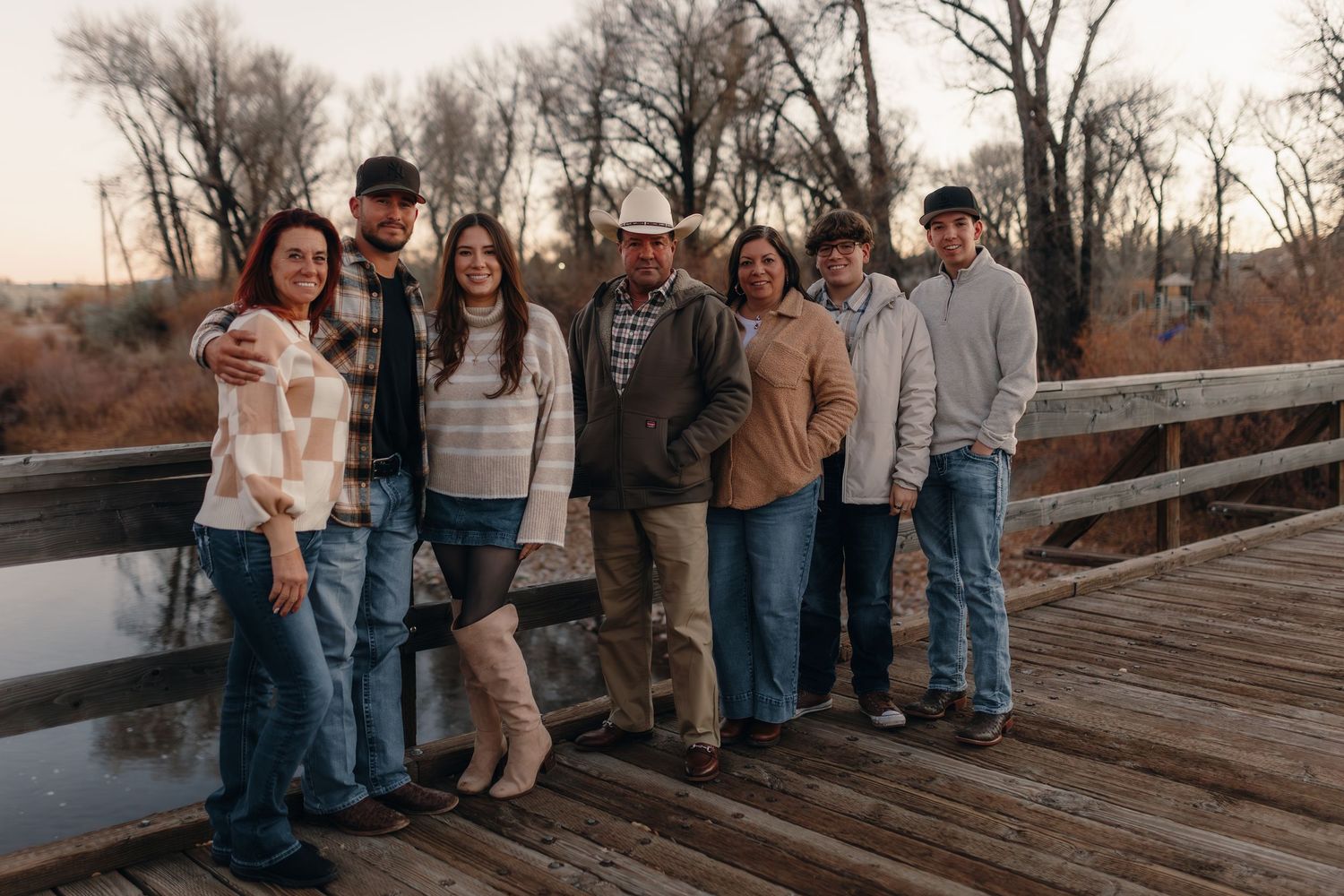 Family of seven posing together on a wooden bridge at dusk, surrounded by bare trees and a calm river.