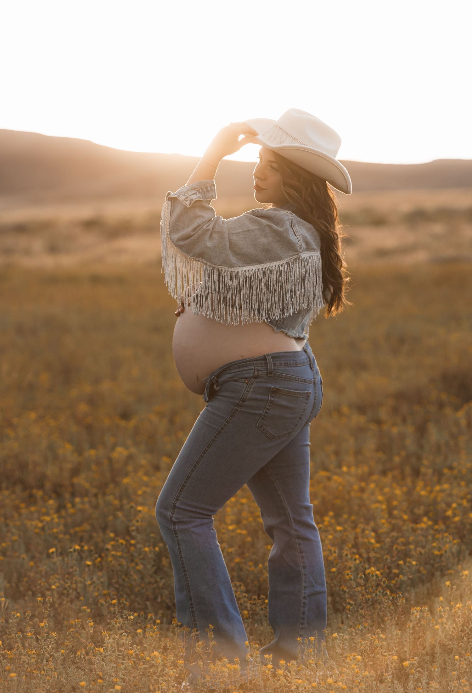 A silhouette of a pregnant woman in western wear against a golden sunset field.