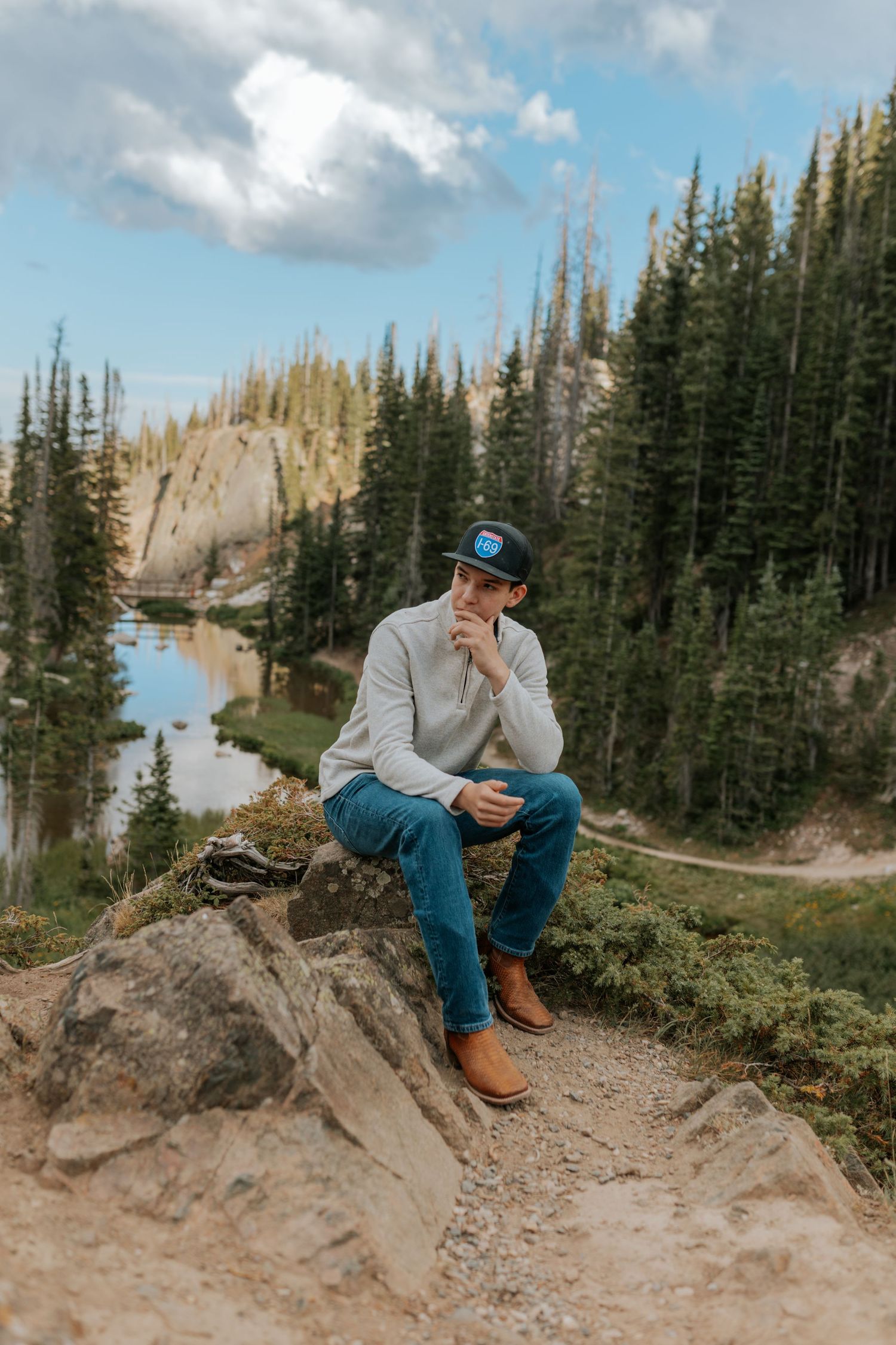 Man in cap and jeans sits on rocky overlook with pine forest and mountain lake backdrop.