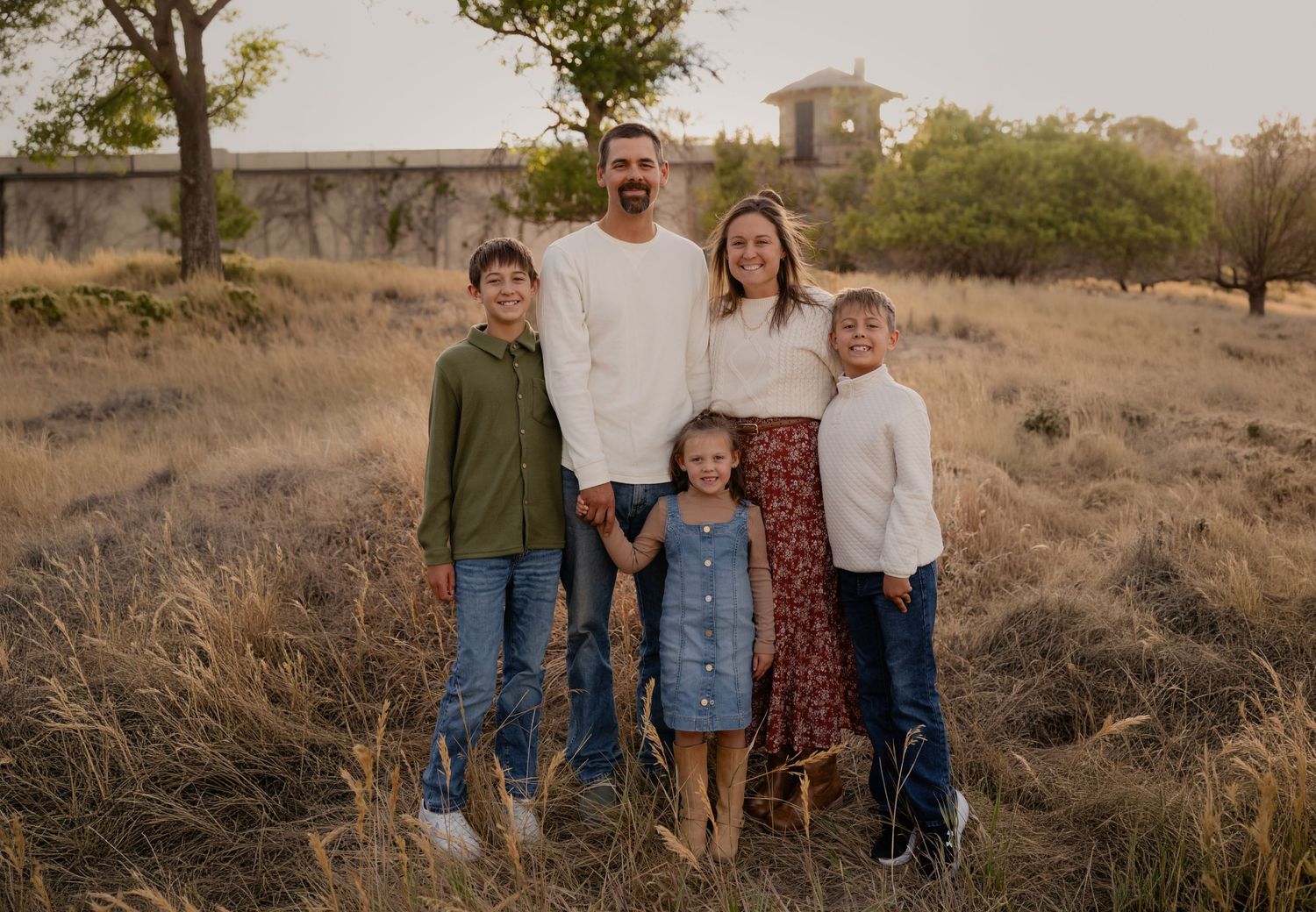 Family of five posing outdoors in a golden field at sunset, parents with three children smiling together.