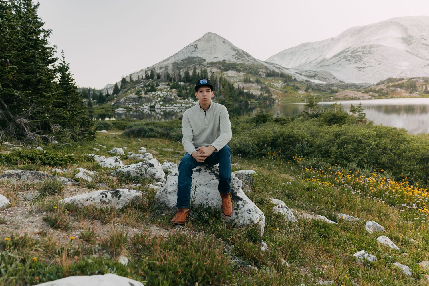 Young man sitting on a boulder in a scenic mountain landscape with a lake and snow-capped peaks in the background.