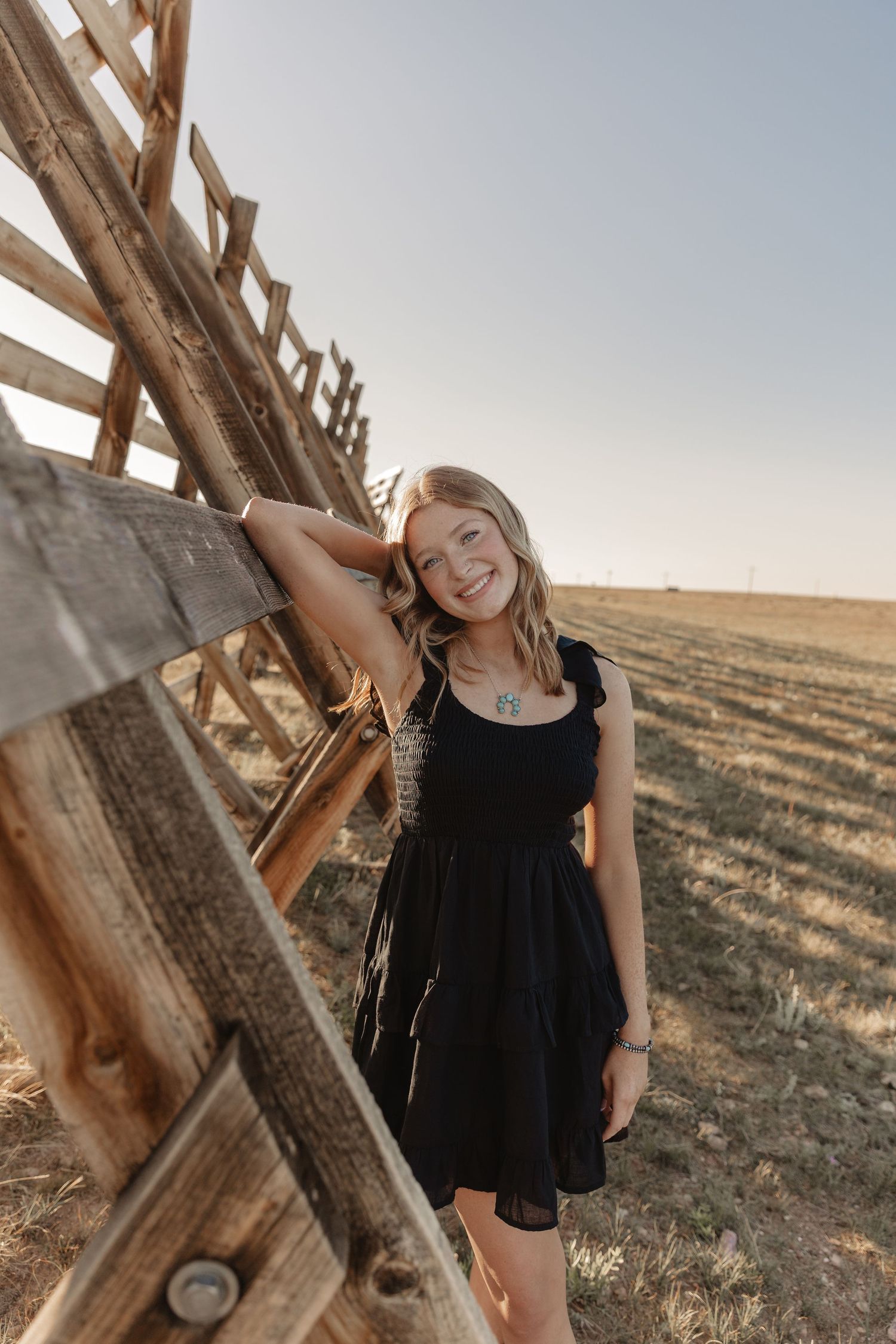 Person in black dress leaning against wooden structure in rural setting at sunset.