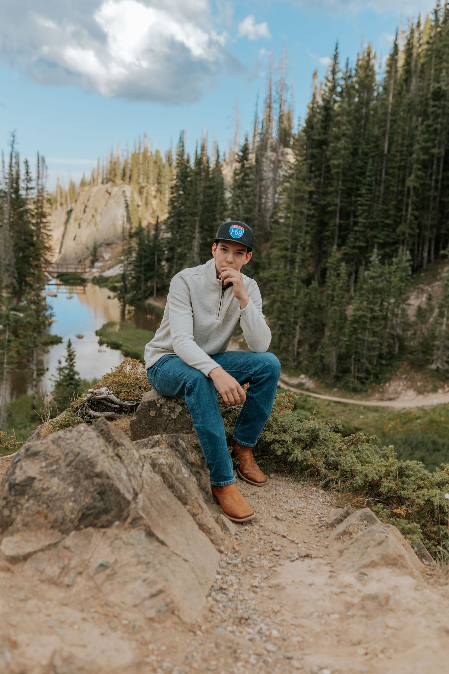 Man in cap and jeans sitting on rocks with mountain lake and pine forest backdrop on a sunny day.
