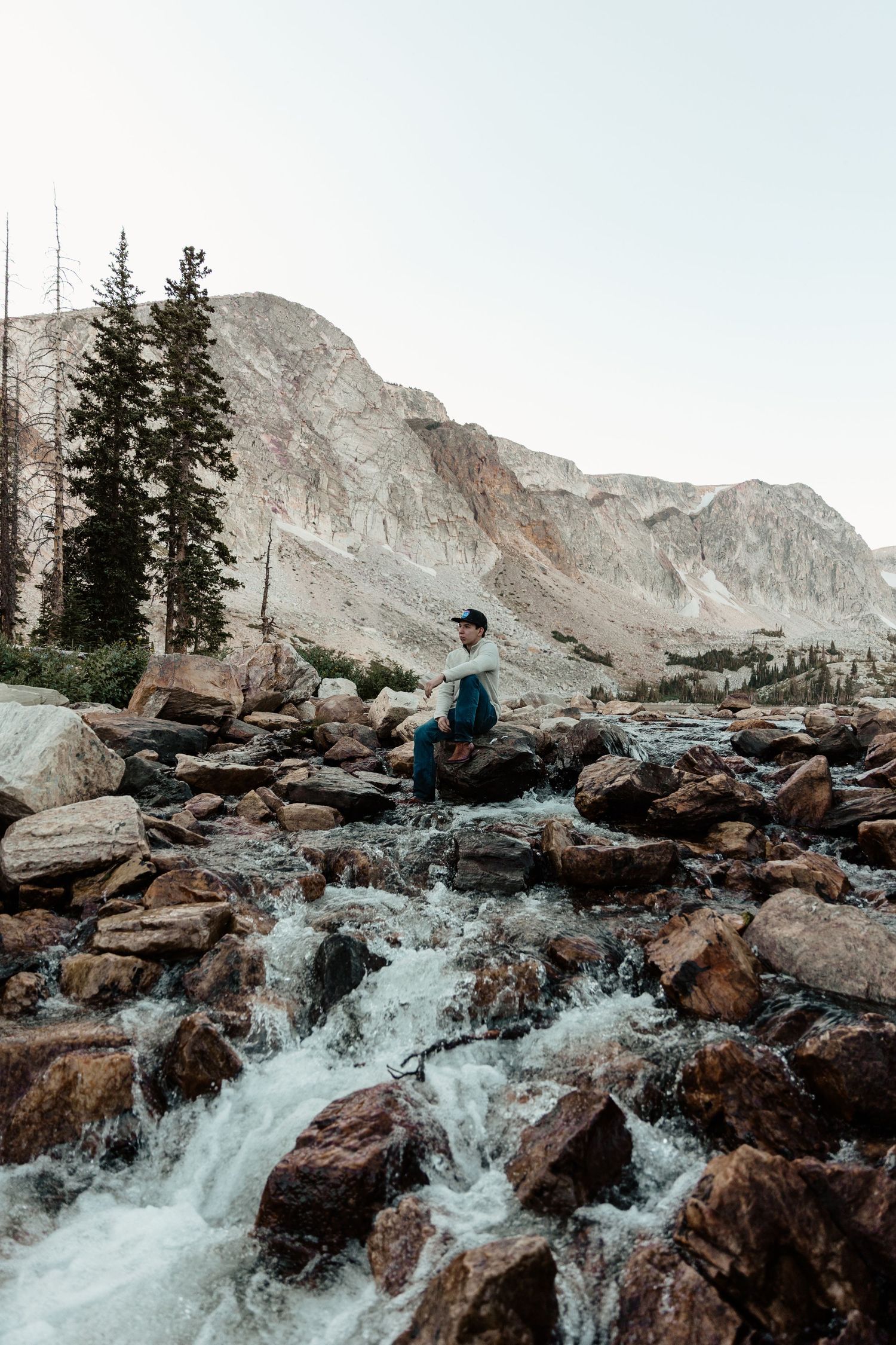 A hiker stands on rocky boulders beside a rushing mountain stream with evergreen trees and peaks behind.