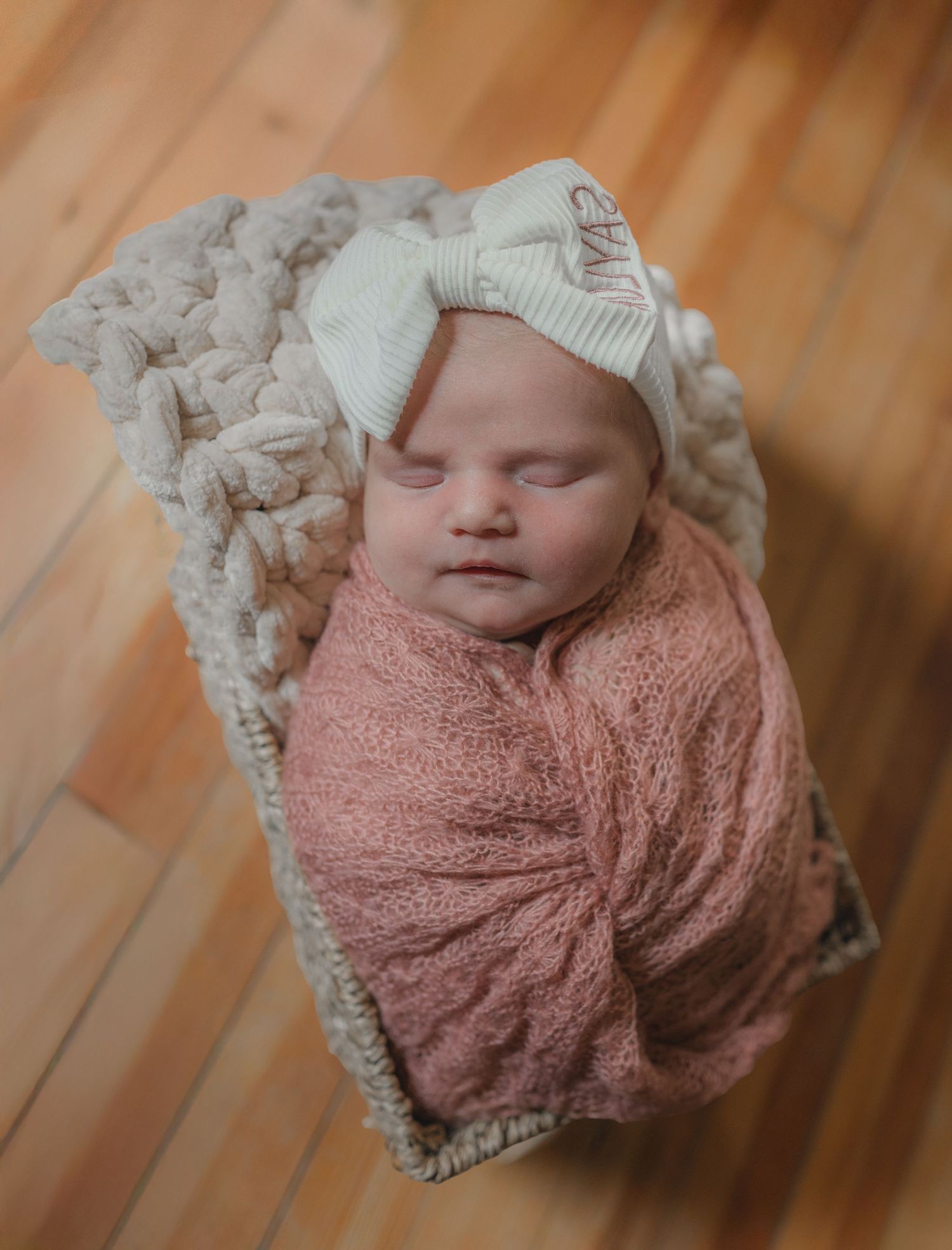 Sleeping newborn wrapped in pink swaddle with white hat resting in textured blanket on wooden floor.