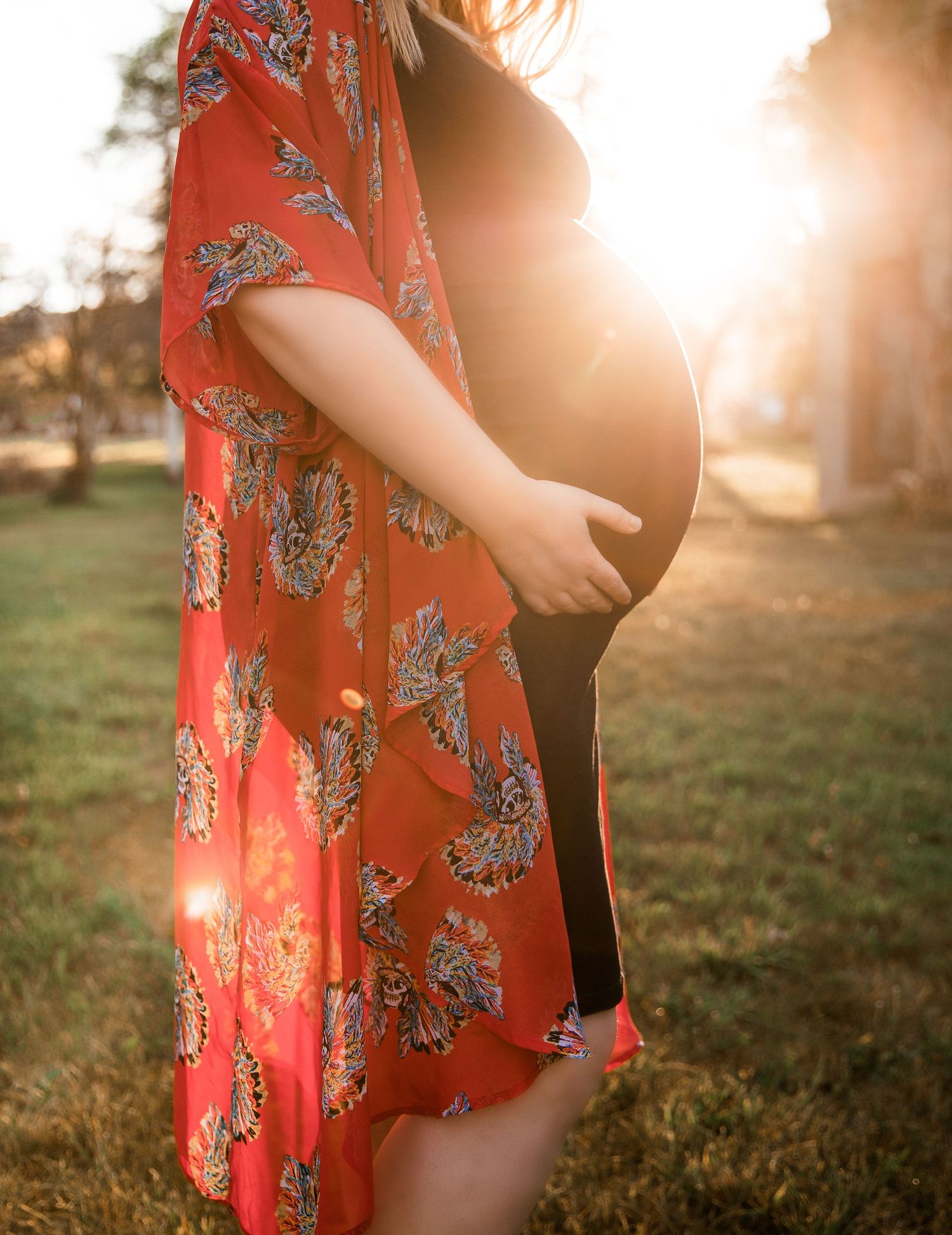 Maternity photo at sunset showing a floral red kimono dress with sunlight creating a warm glow in the background.