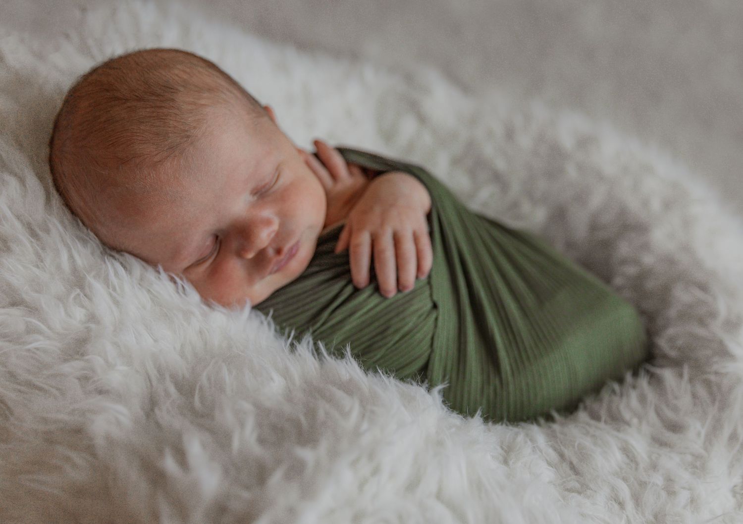 Newborn wrapped in olive green fabric sleeping peacefully on white fur rug.