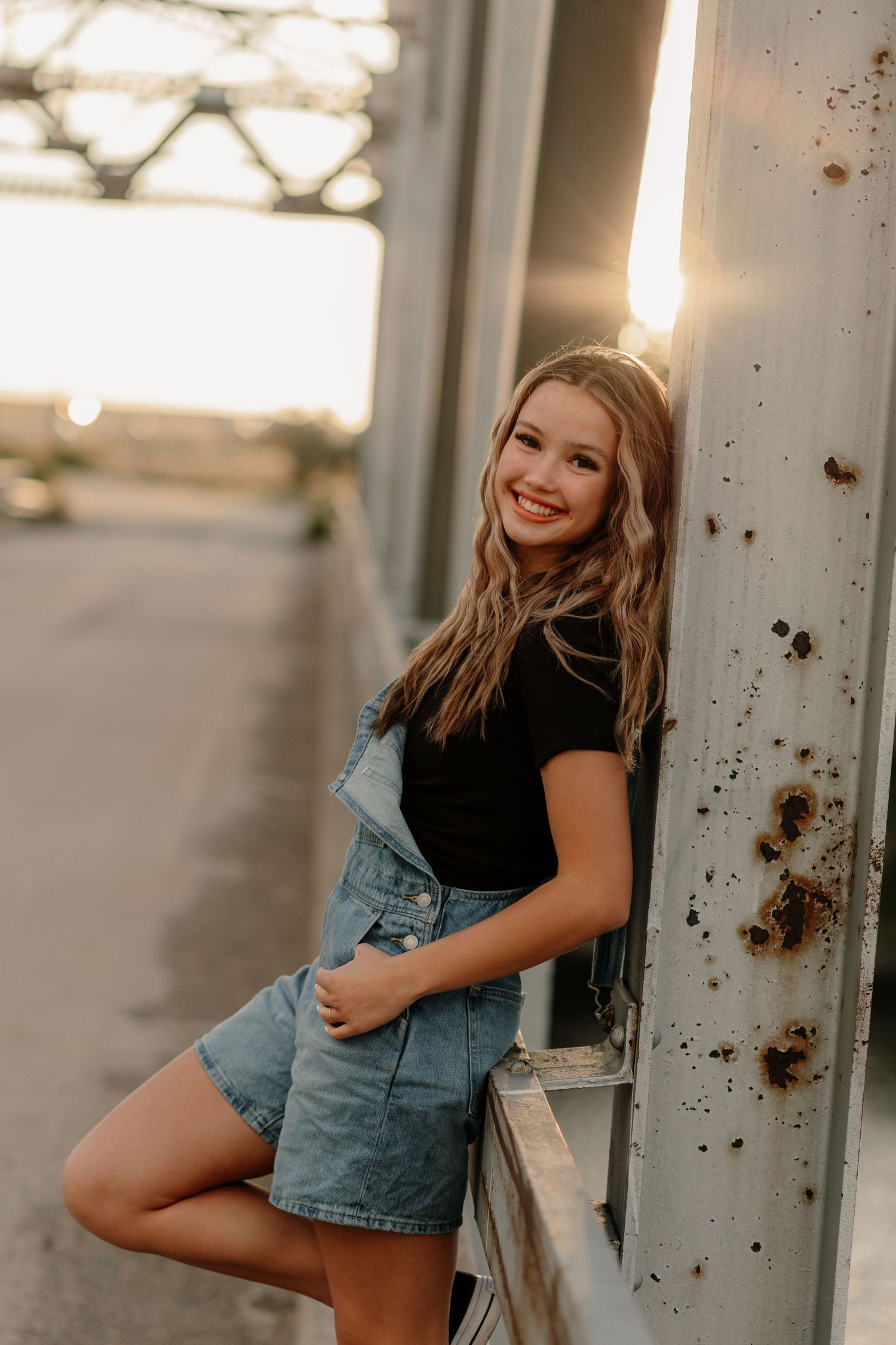 Smiling teenage girl in denim overalls and black top leaning against rusty metal pillar at golden hour.