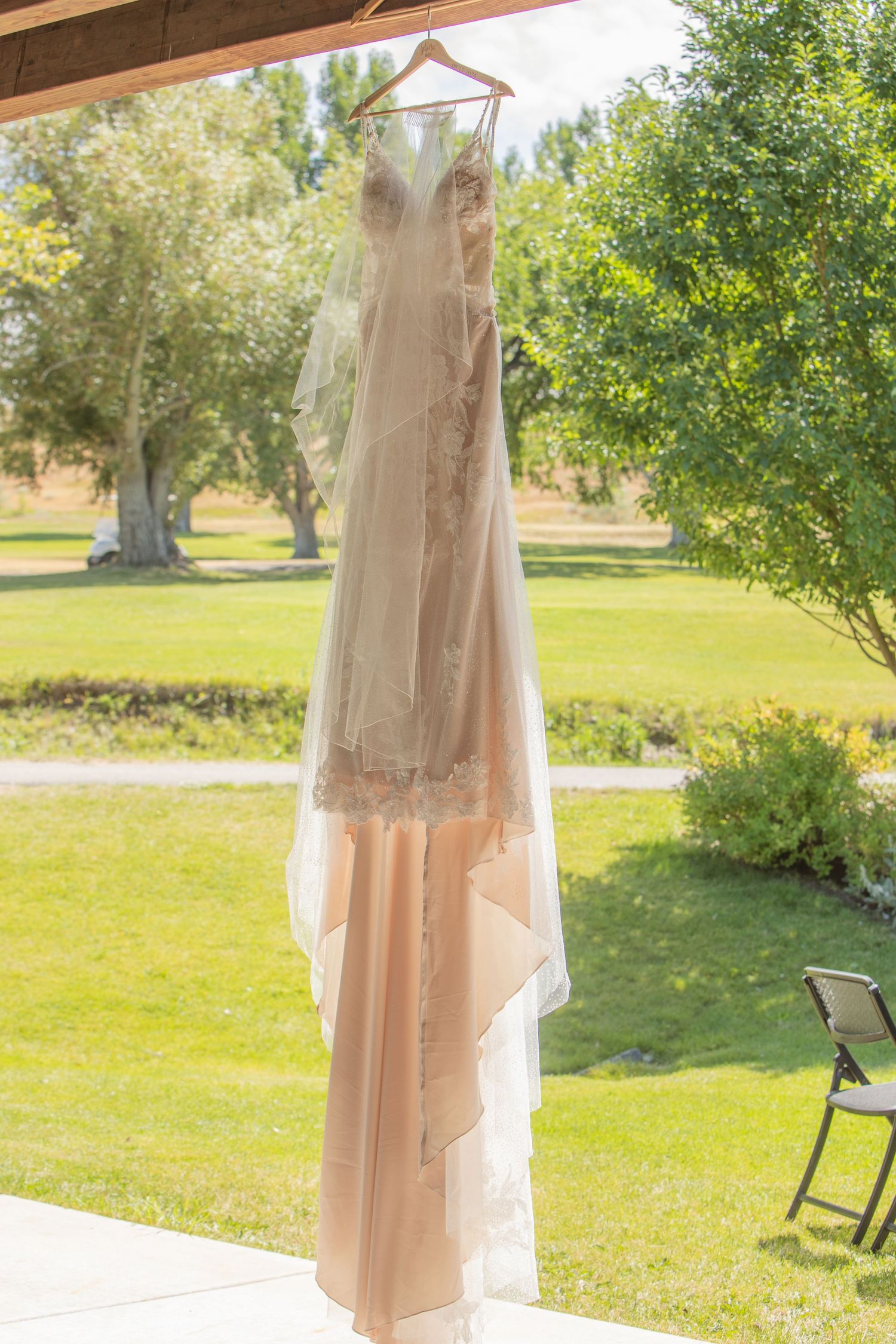 An elegant wedding dress with veil hangs from a copper-colored hanger outdoors against a sunlit garden background.