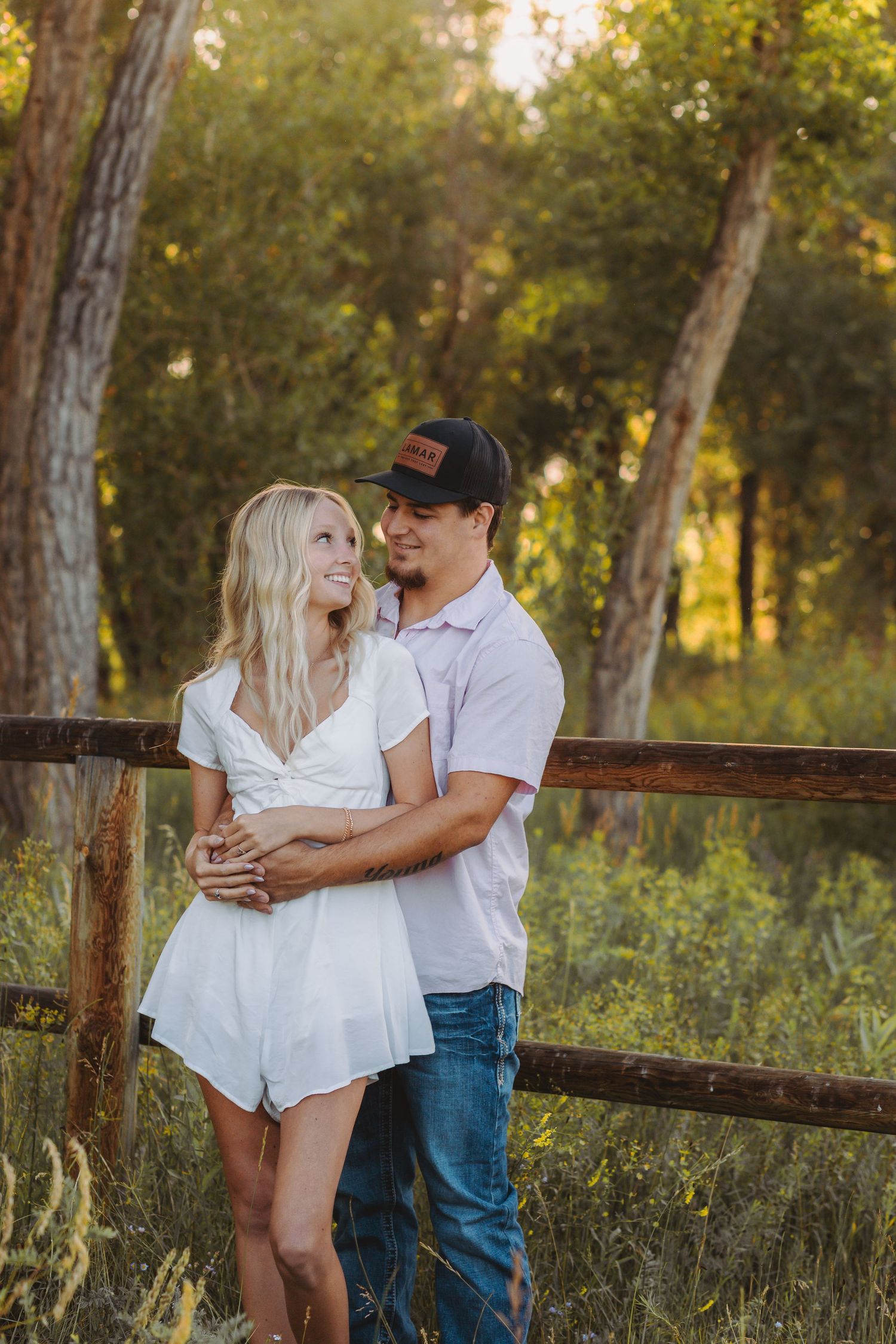 Couple embrace near rustic wooden fence in sunlit forest setting during golden hour.