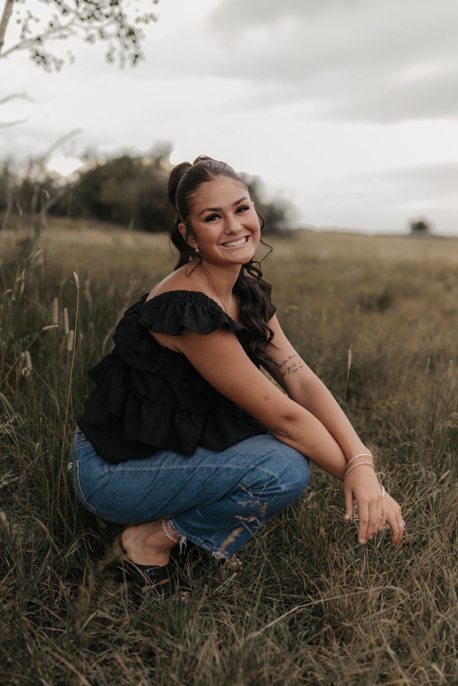 Young woman in black off-shoulder top and jeans crouching in a grassy field, smiling outdoors.
