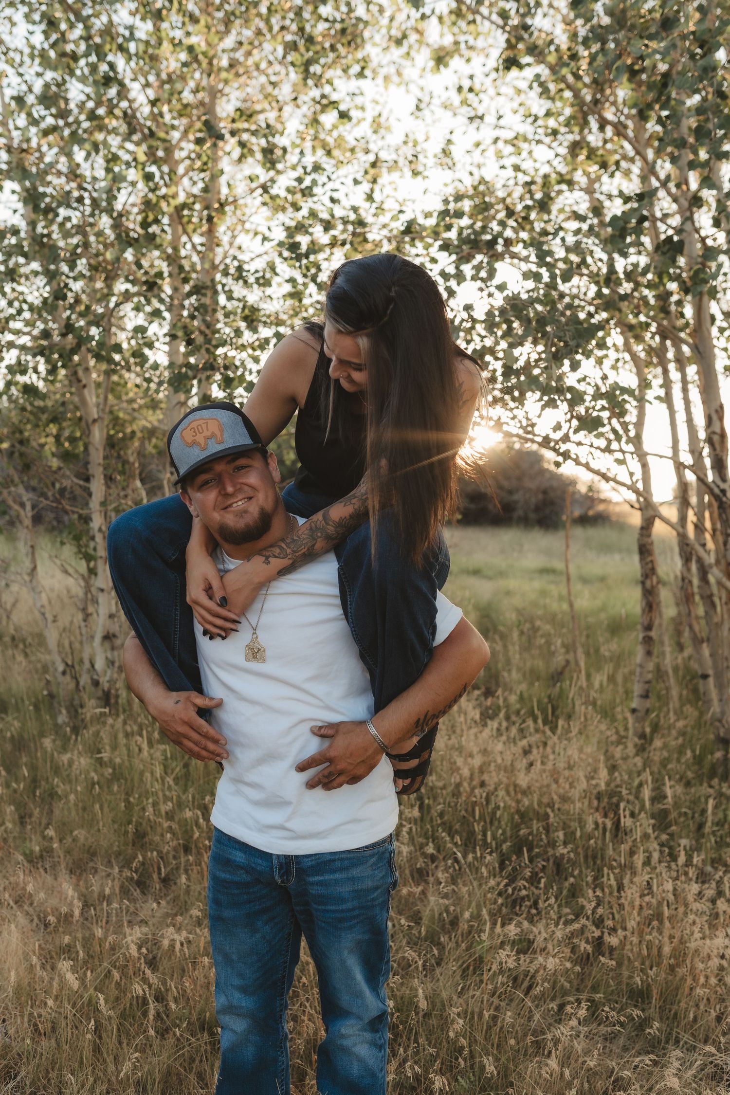 A couple shares a piggyback ride in a sunlit birch forest, the woman hugging the man from behind as golden light filters through trees.