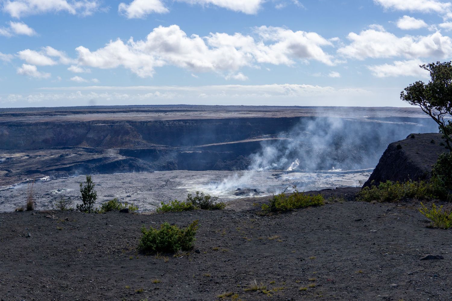Steam rises from the Kilauea Volcano crater in Hawaii Volcanoes National Park against a blue sky with white clouds.