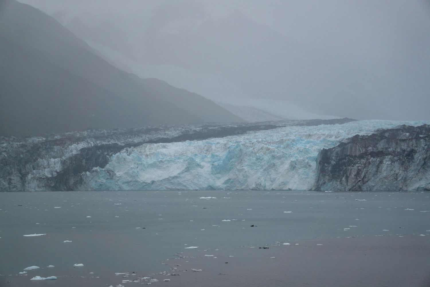 Massive blue glacier face meets the ocean with floating ice chunks in misty conditions.