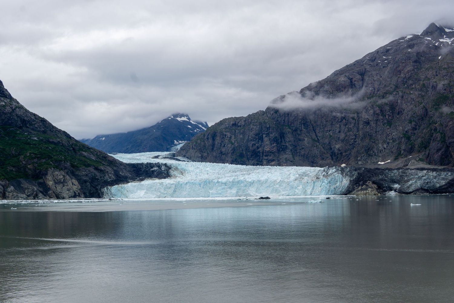 A majestic tidewater glacier terminus meets the waters of an Alaskan fjord beneath cloudy skies.