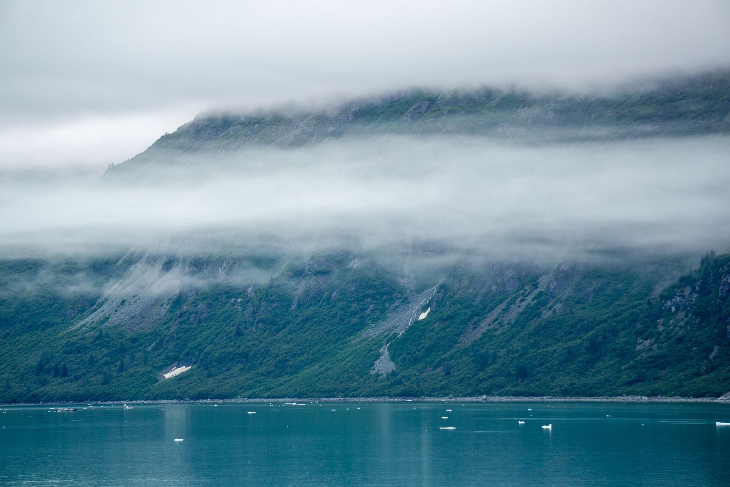 Misty mountains meet turquoise waters in a dramatic fjord landscape with low-hanging clouds.