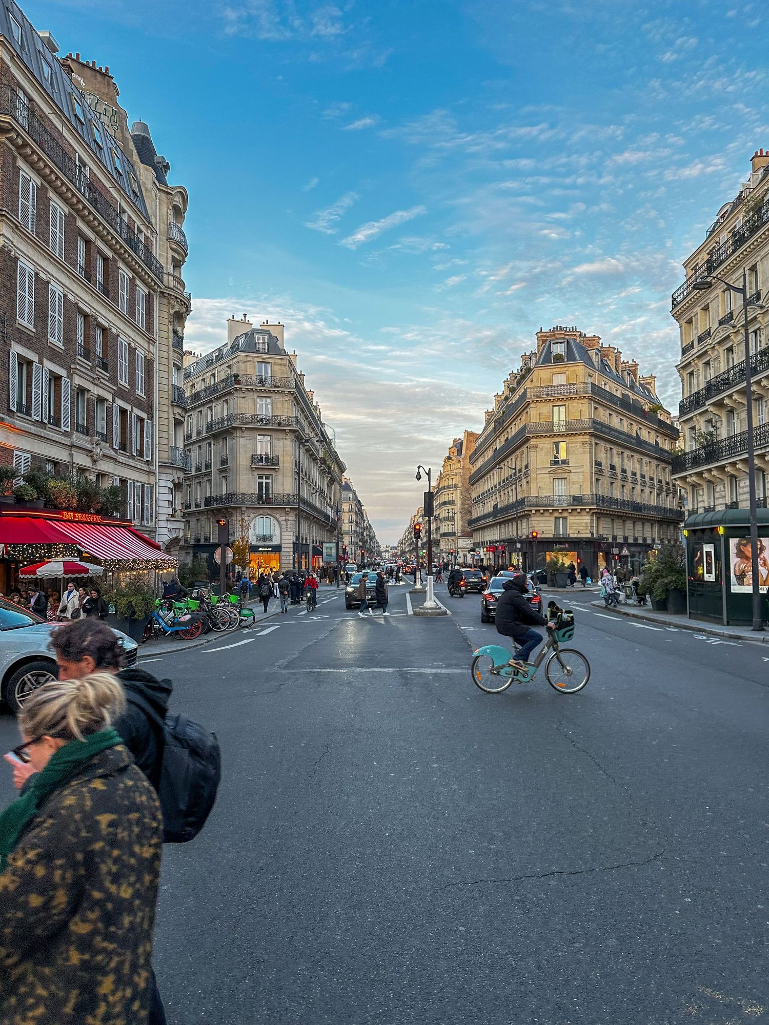 A scenic Parisian intersection with classic Haussmann architecture, cyclists, and a vibrant street scene under a blue sky.
