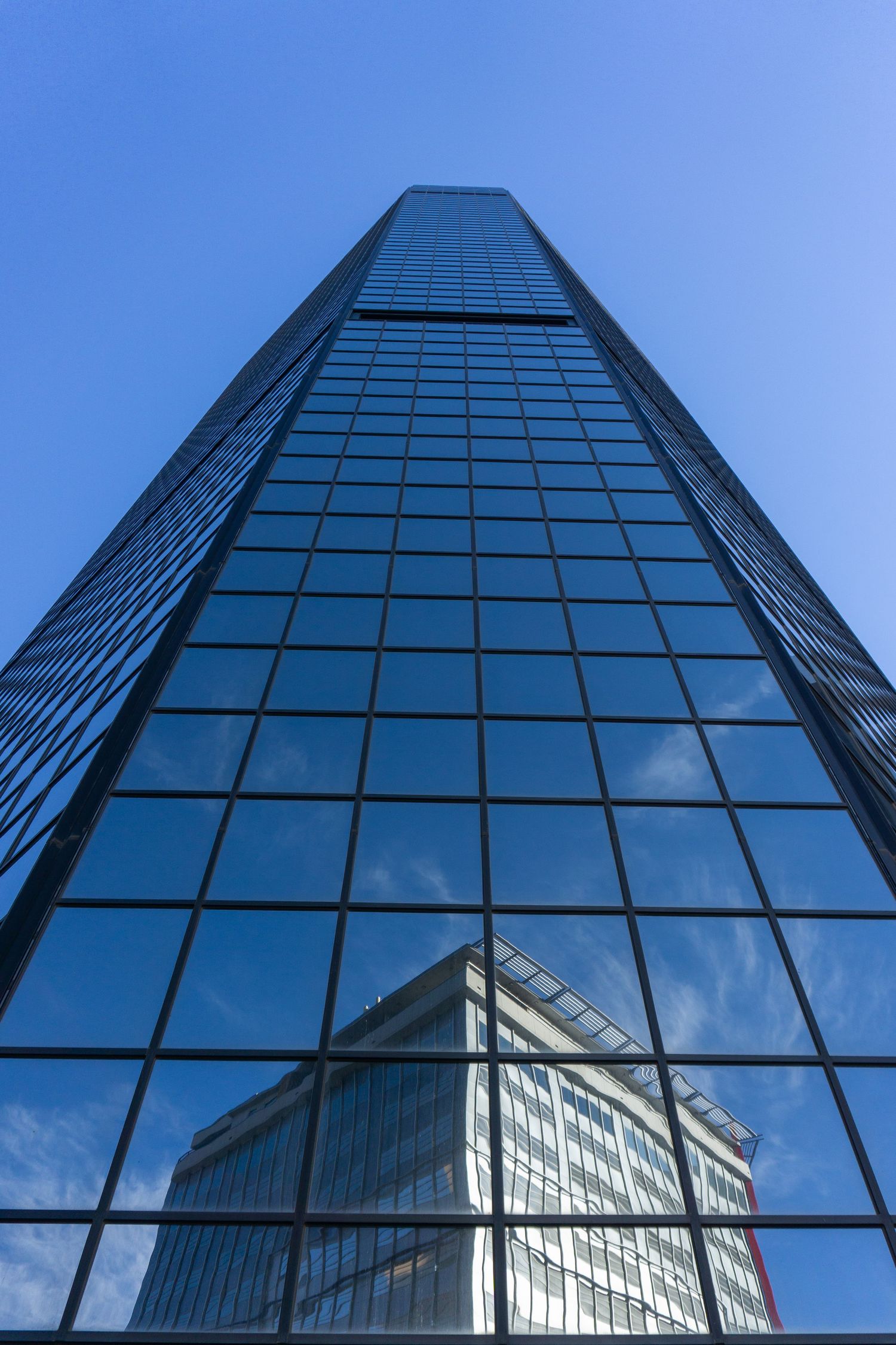 Modern glass skyscraper photographed from ground level against bright blue sky reflecting clouds.