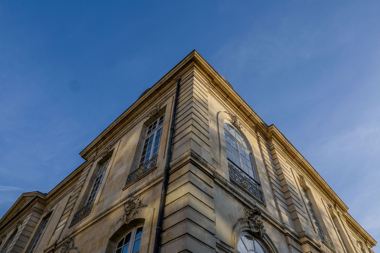 Low angle view of an ornate historic beige building facade with arched windows against a bright blue sky.
