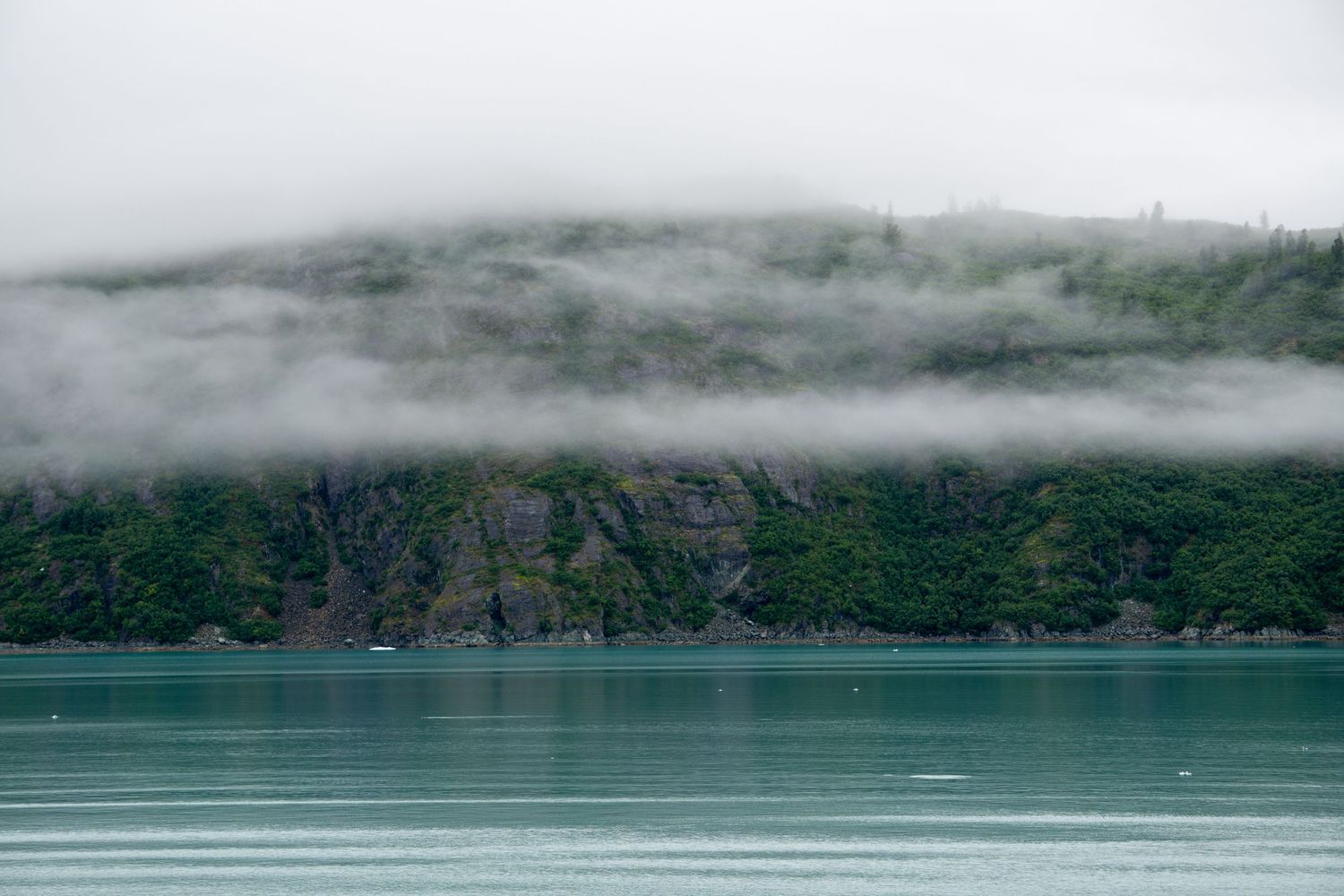Fog rolls over rocky cliffs and evergreen forest along turquoise glacial waters.