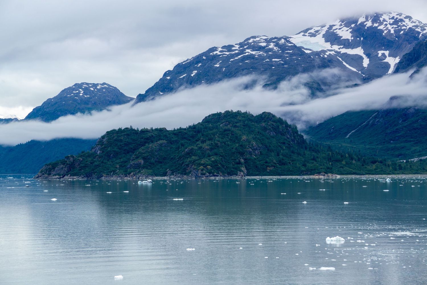 Low clouds wrap around coastal mountains above a bay scattered with small icebergs.